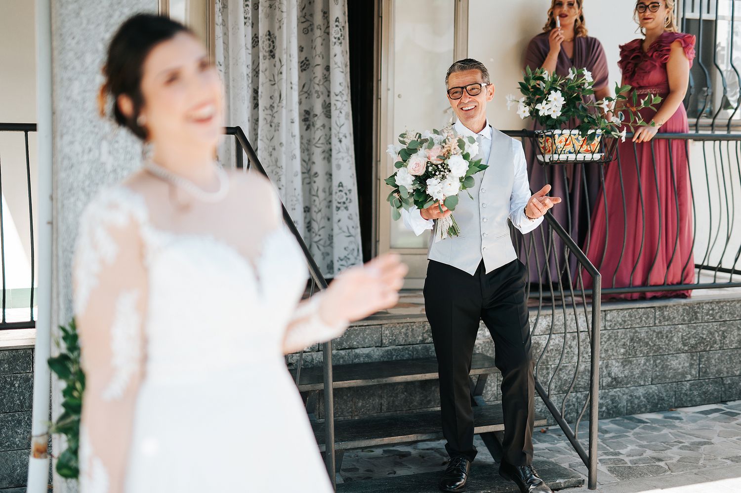 Groom holding bouquet smiling at bride during outdoor wedding ceremony on steps.