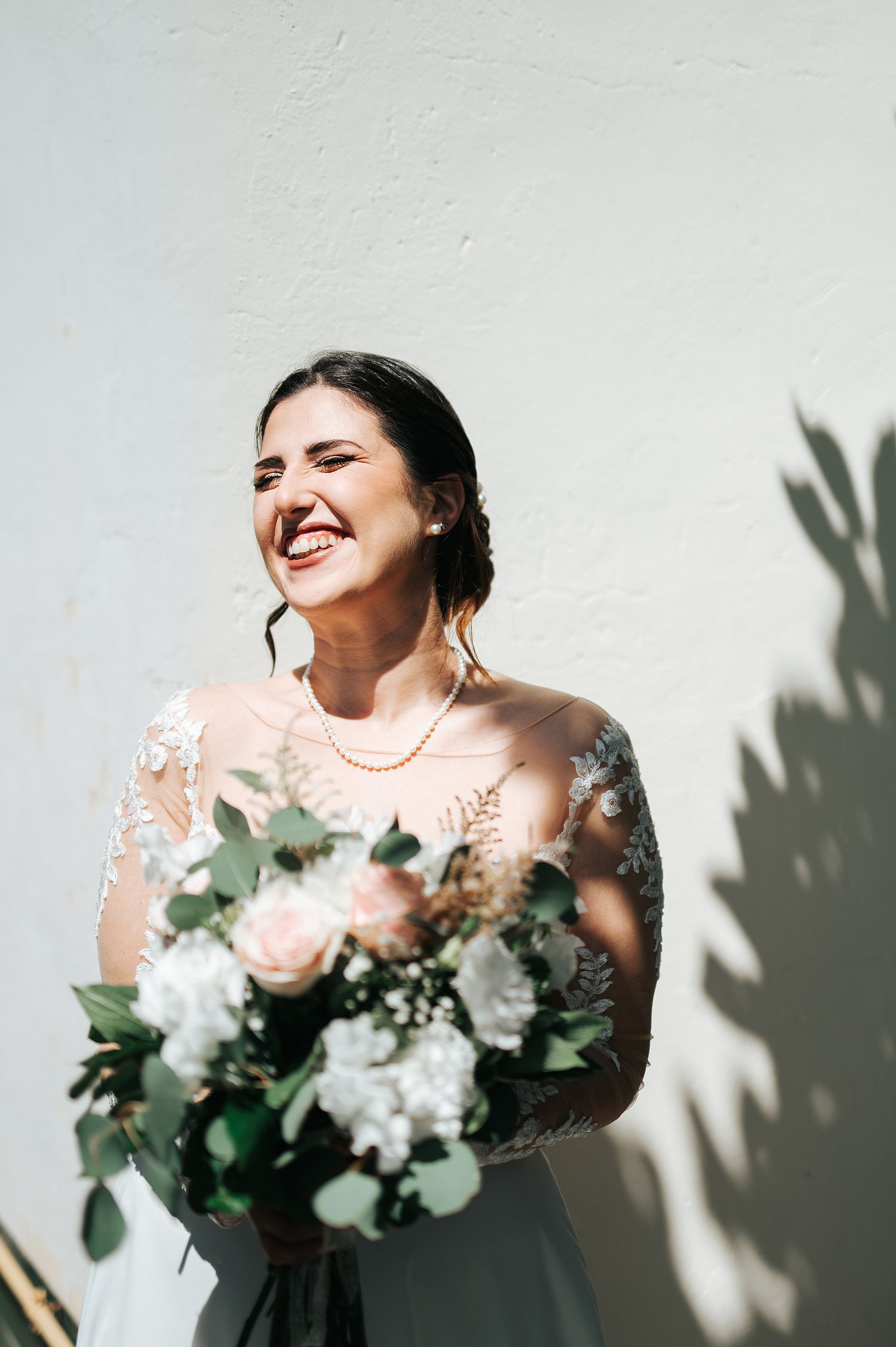 Happy bride in lace gown holding pink and white bouquet against white wall.