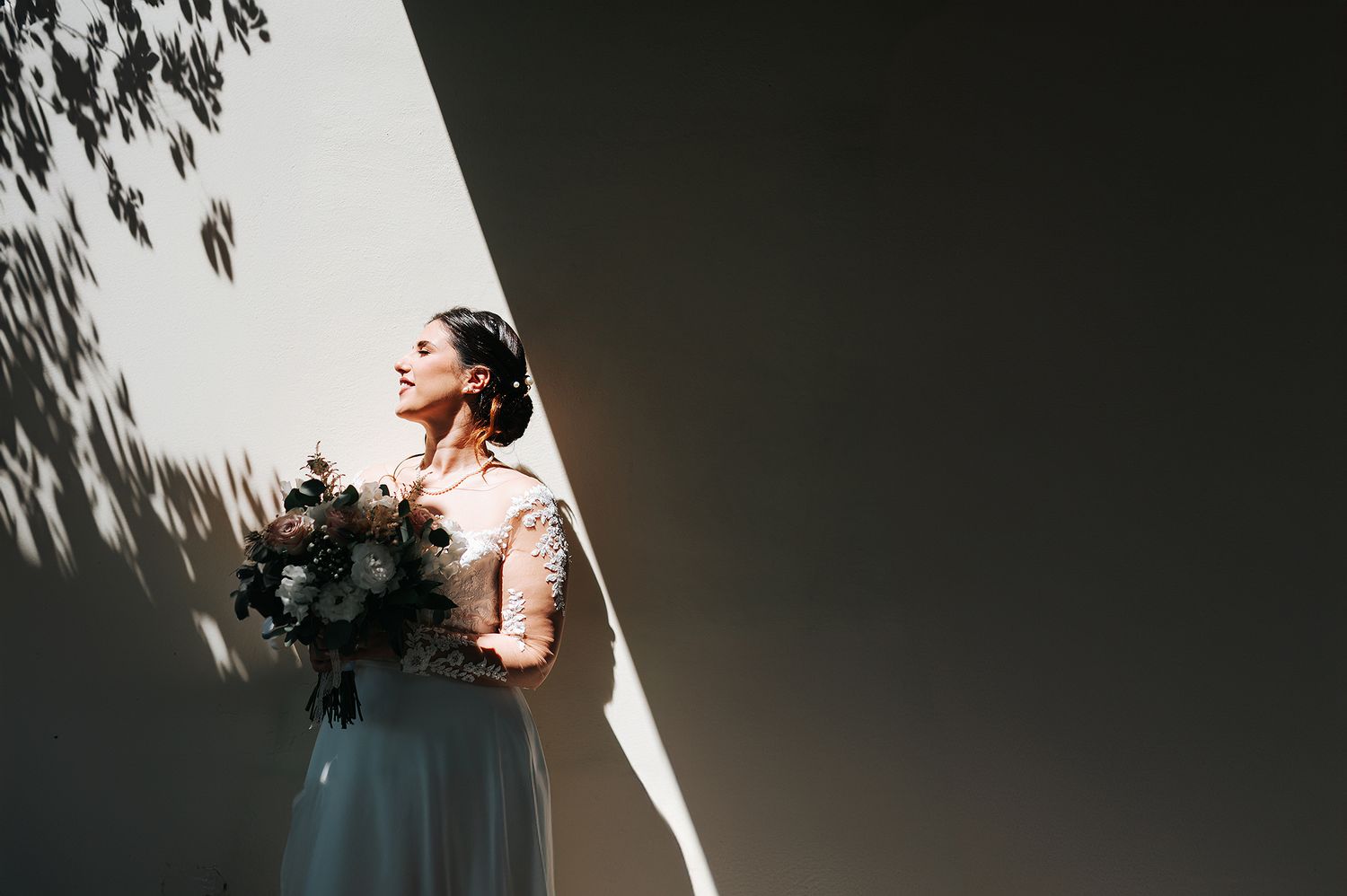 Radiant bride holding bouquet standing in dramatic sunlight against white wall.