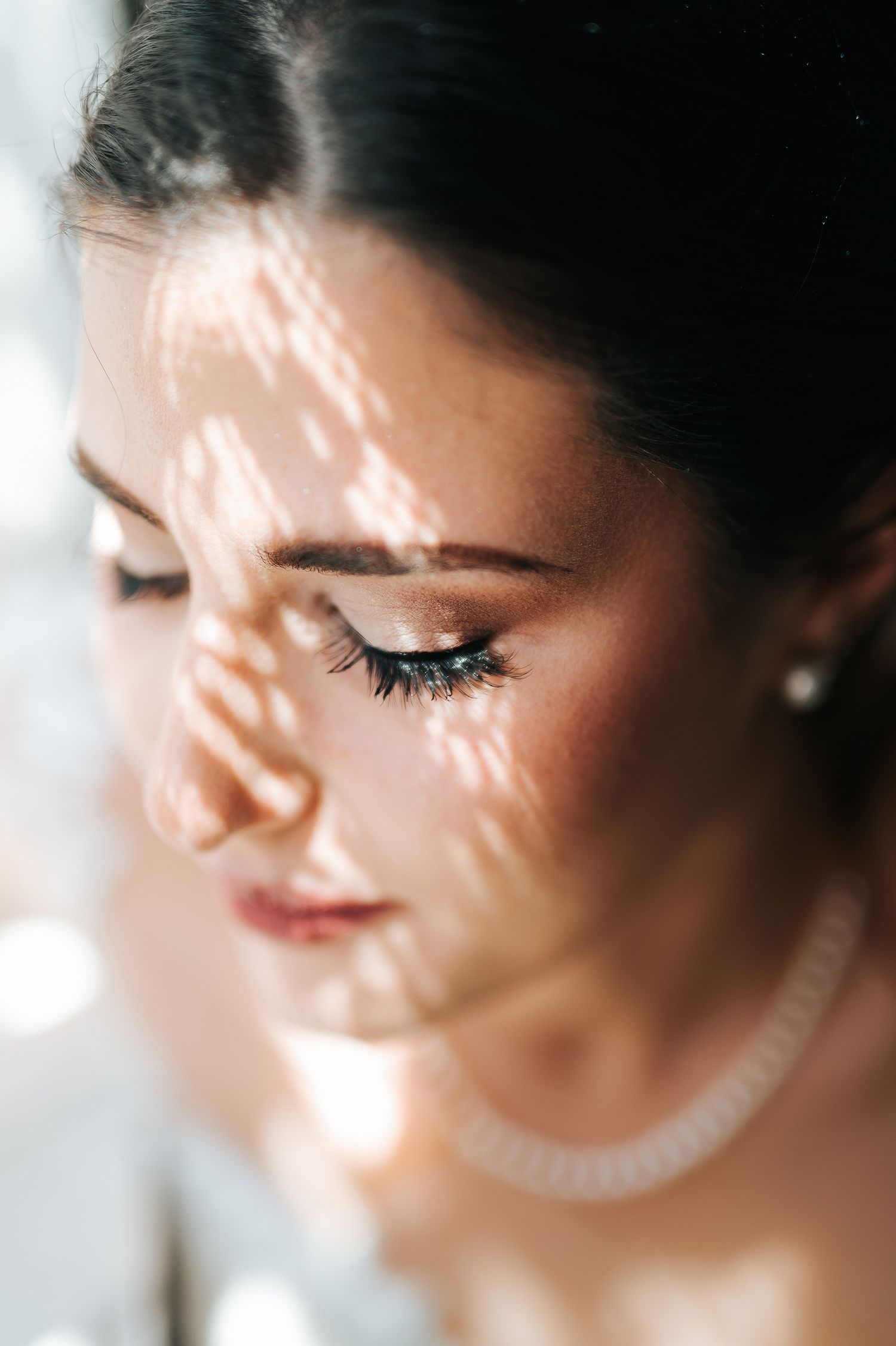 Close-up of a bride with closed eyes, wearing pearl earrings and necklace, with dappled sunlight on her face.