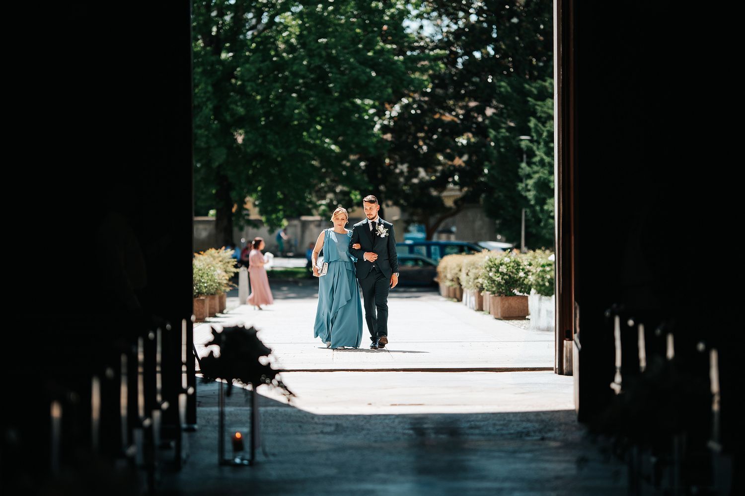 Couple walking through grand doors into sunlit courtyard surrounded by lush green trees at wedding venue.