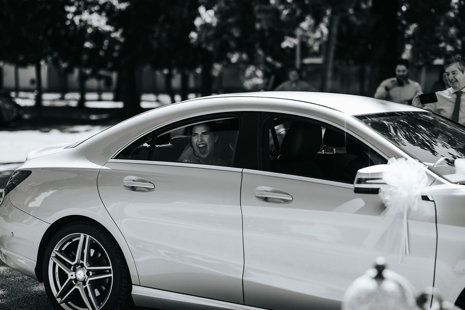 Black and white photo of smiling bride seen through window of decorated white Mercedes wedding car.