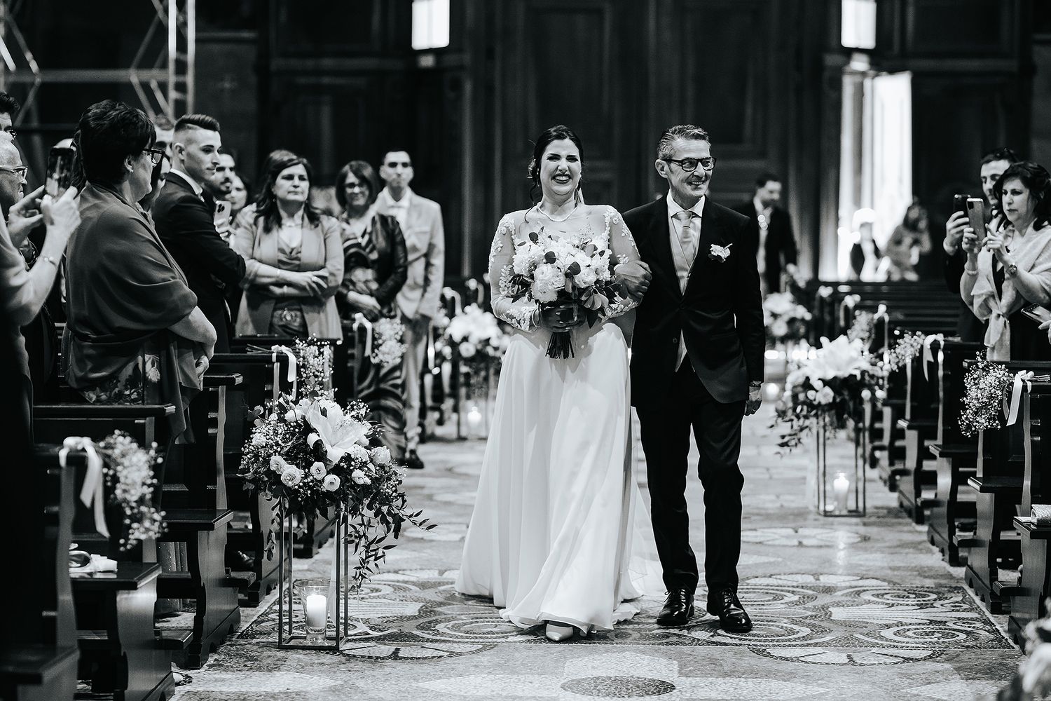 Black and white photo of bride walking down church aisle with escort, flanked by floral arrangements.