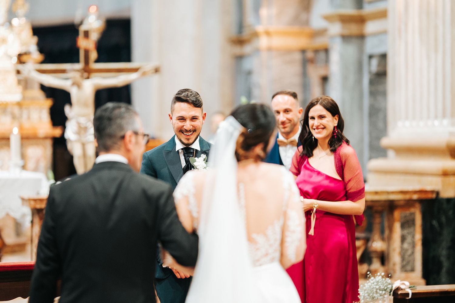 Groom smiling joyfully as bride approaches altar in ornate church, with wedding party guests behind.