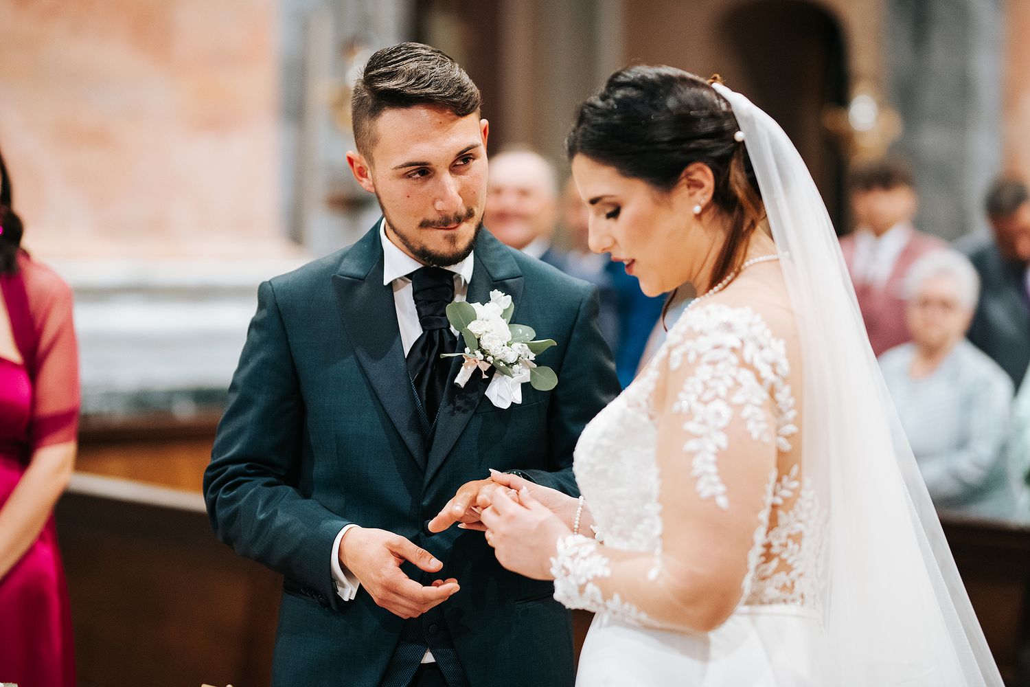 Groom placing wedding ring on bride's finger during church ceremony, both dressed elegantly.