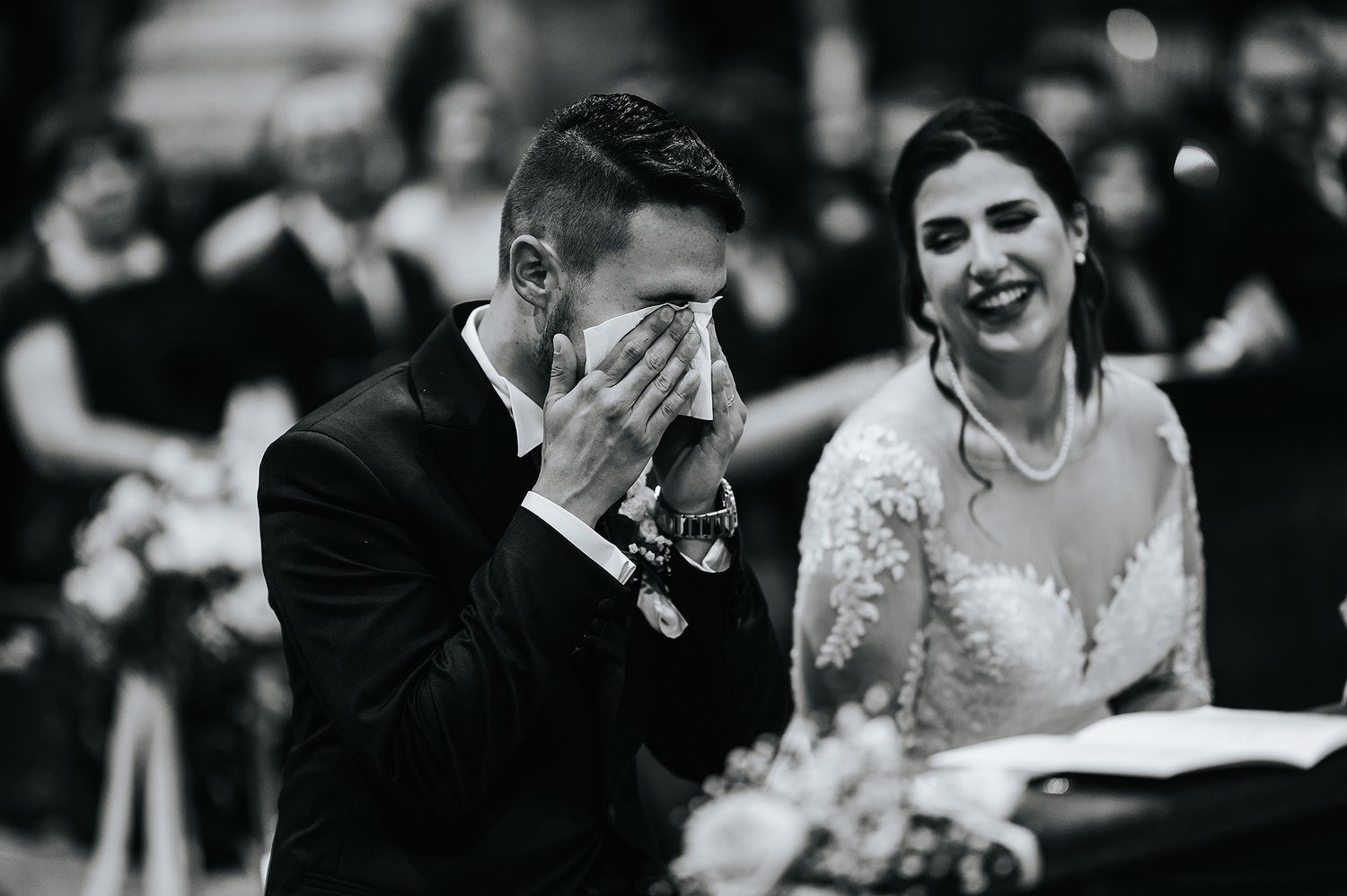Emotional groom covering face with hands while bride smiles during wedding ceremony, black and white.