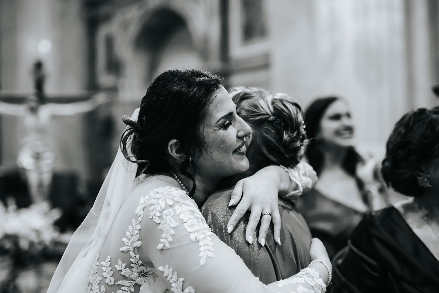 Bride in lace wedding dress sharing heartfelt hug with guest during wedding ceremony.