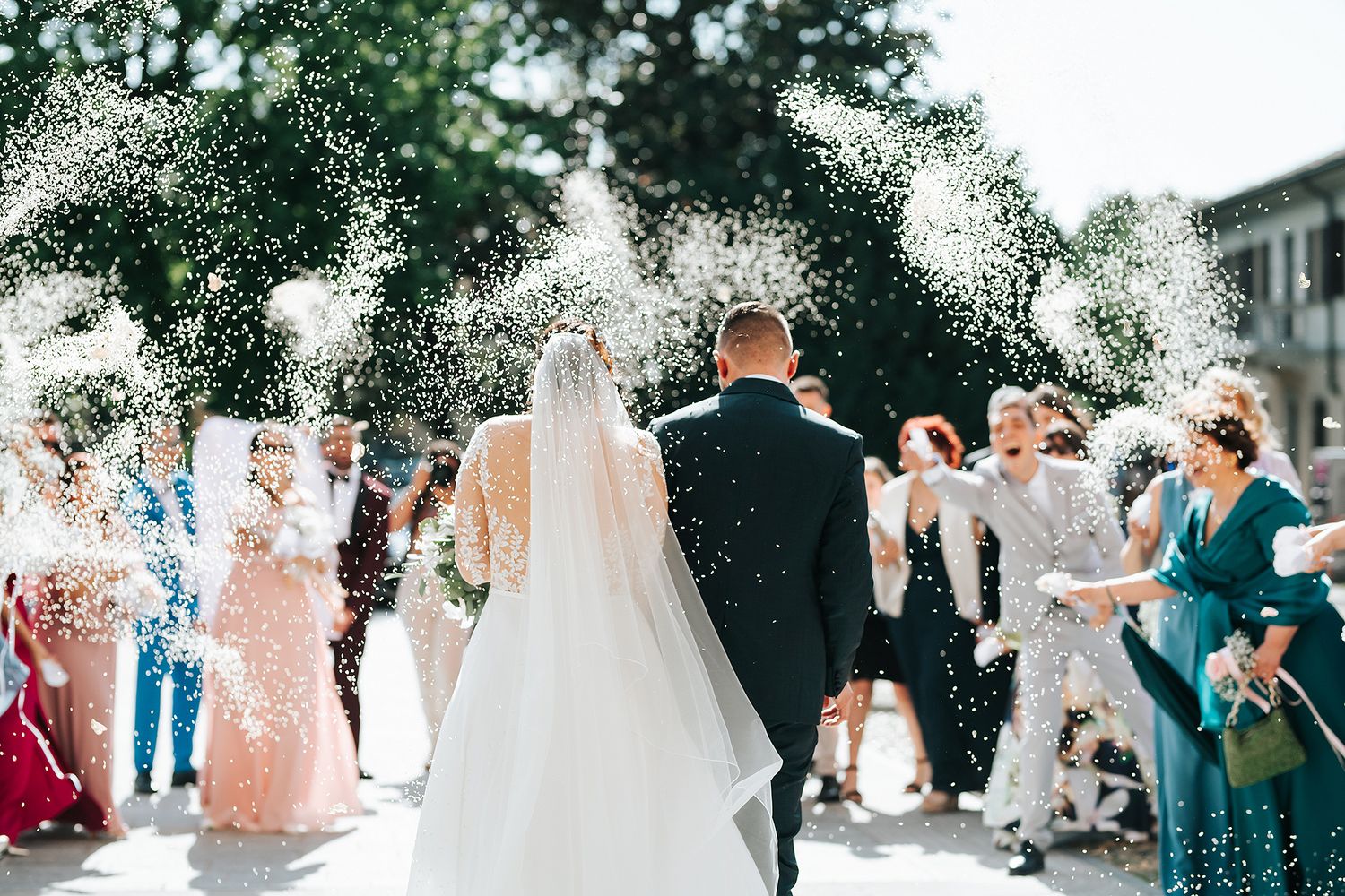 Newlyweds walking through confetti shower thrown by cheering wedding guests outdoors.