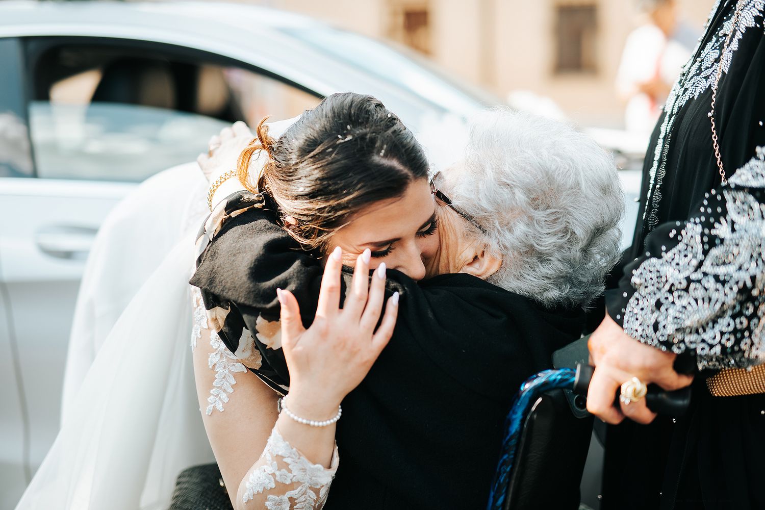 Emotional bride in lace gown embracing elderly woman in wheelchair after wedding ceremony.