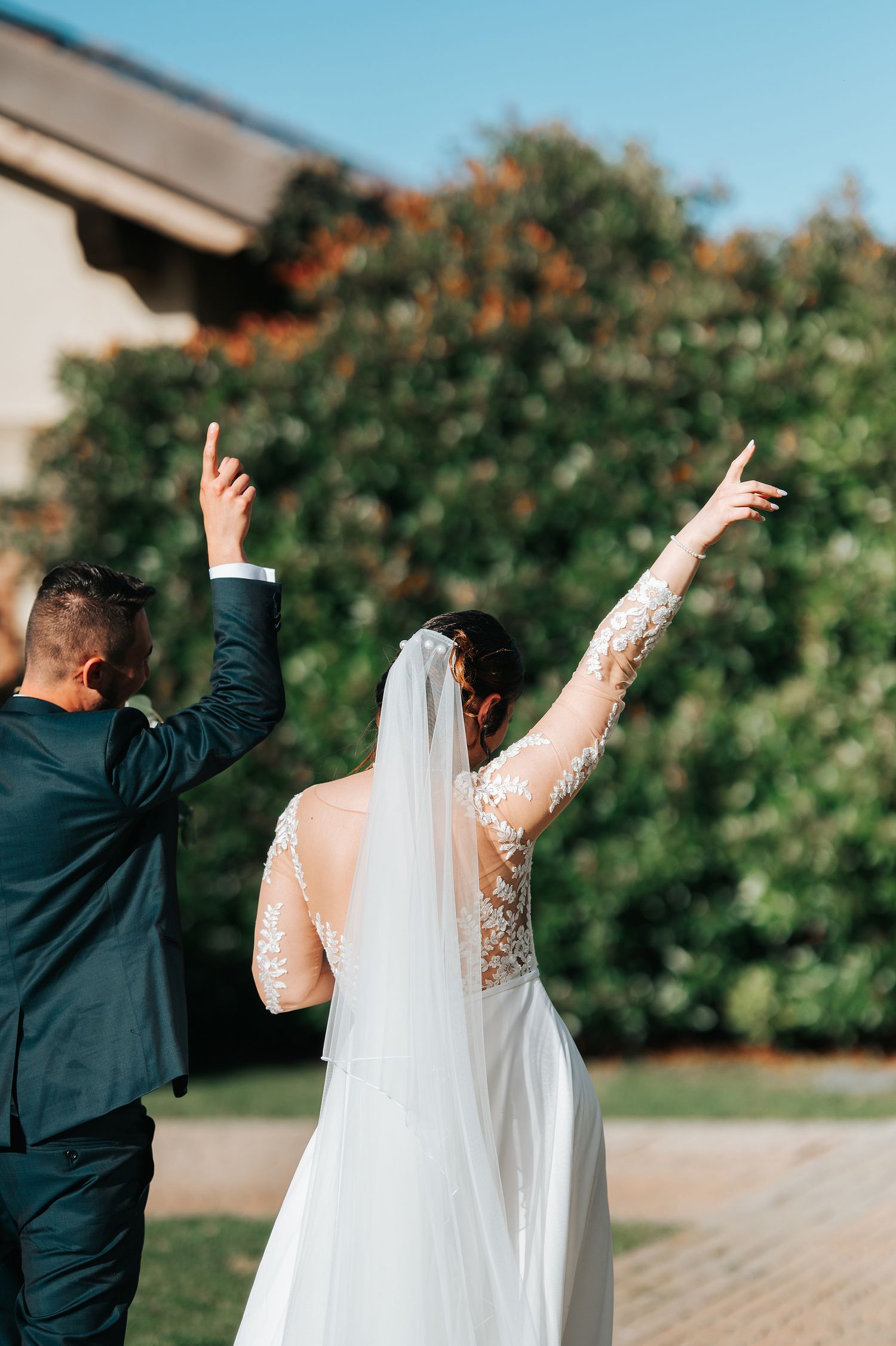 Bride and groom raising arms in celebration outdoors with lush green hedges behind them.