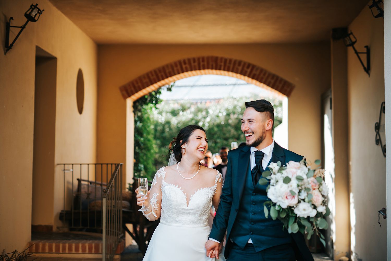 Joyful newlyweds walking hand in hand under rustic archway, bride holding bouquet.