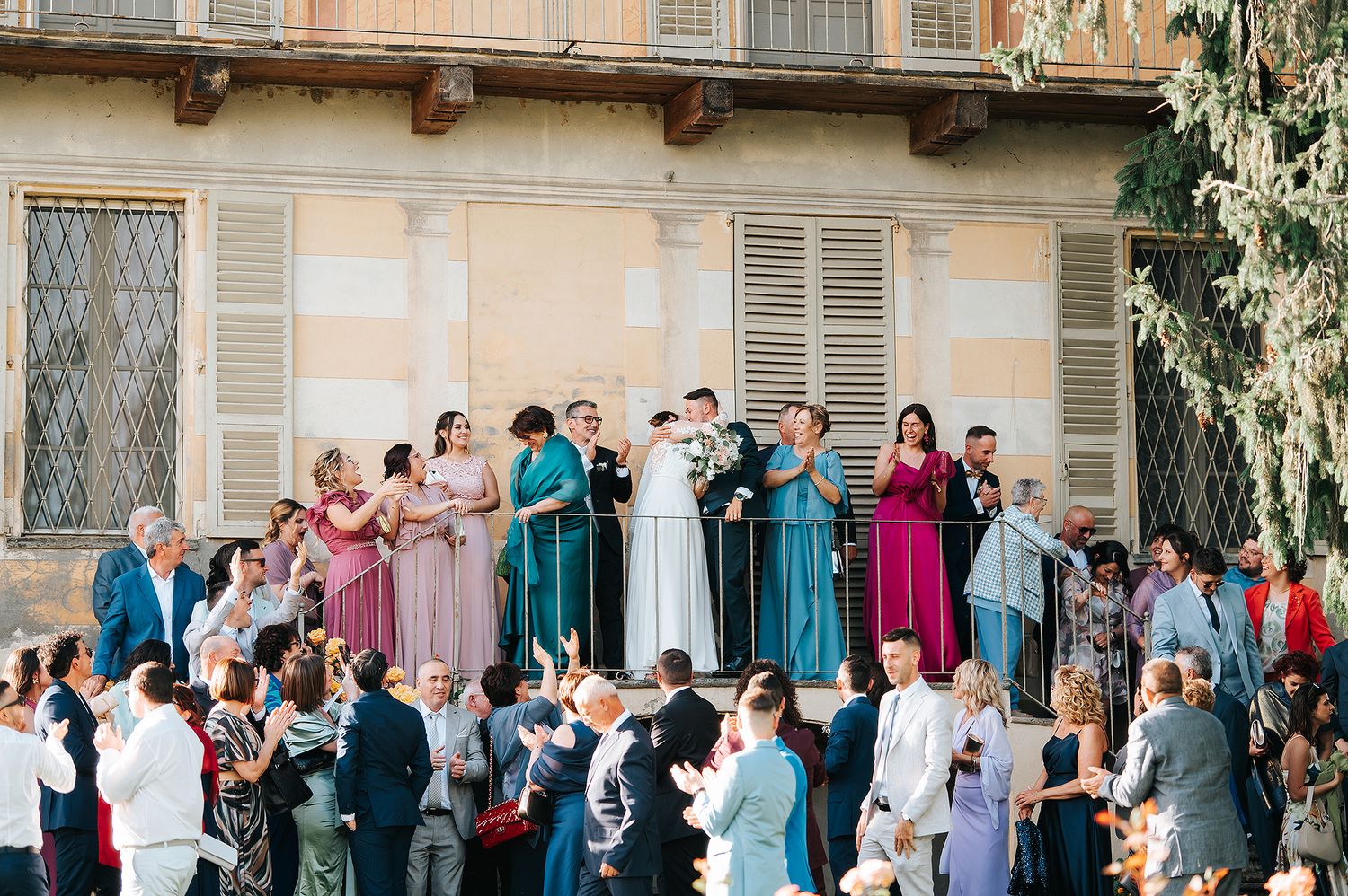 Wedding party and guests celebrate on a villa balcony in a sunny Italian courtyard.