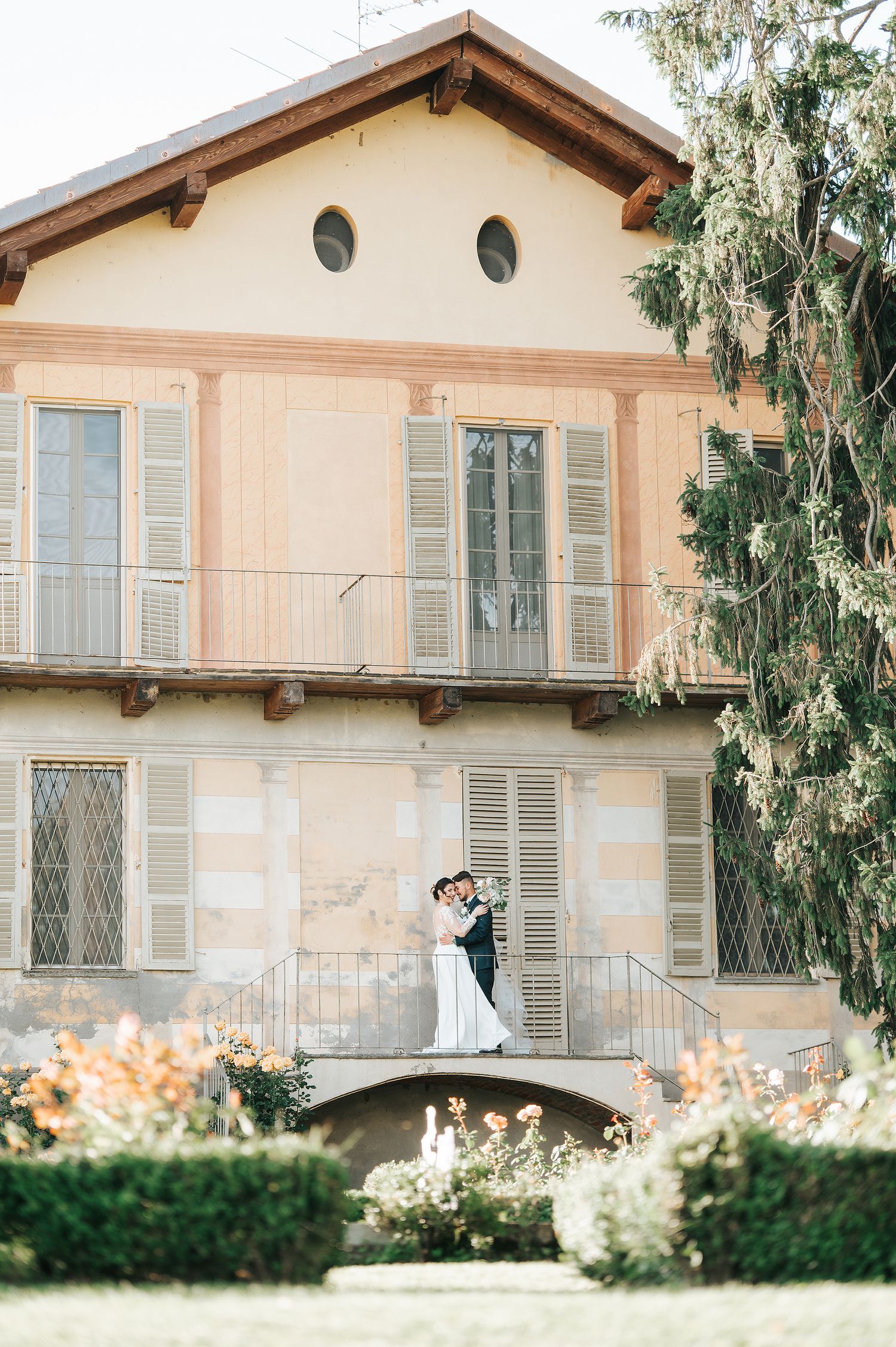Newlyweds embrace on a balcony of a beautiful Italian villa surrounded by gardens.
