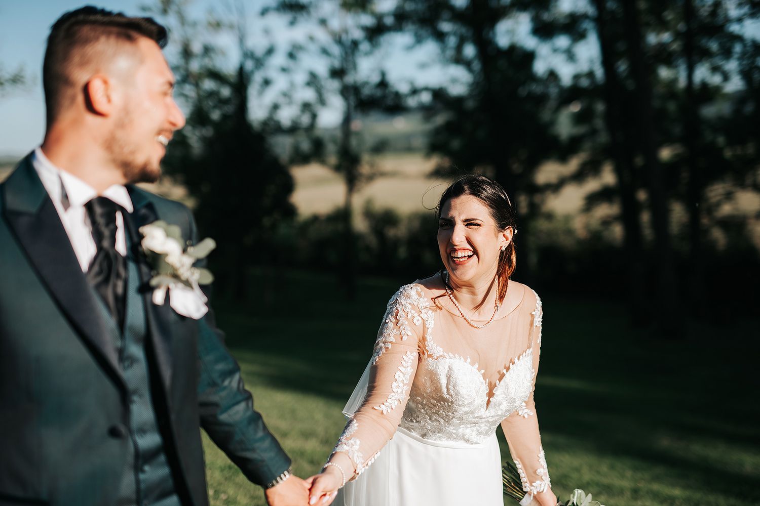 Smiling bride in lace gown holds grooms hand outdoors in golden afternoon light.