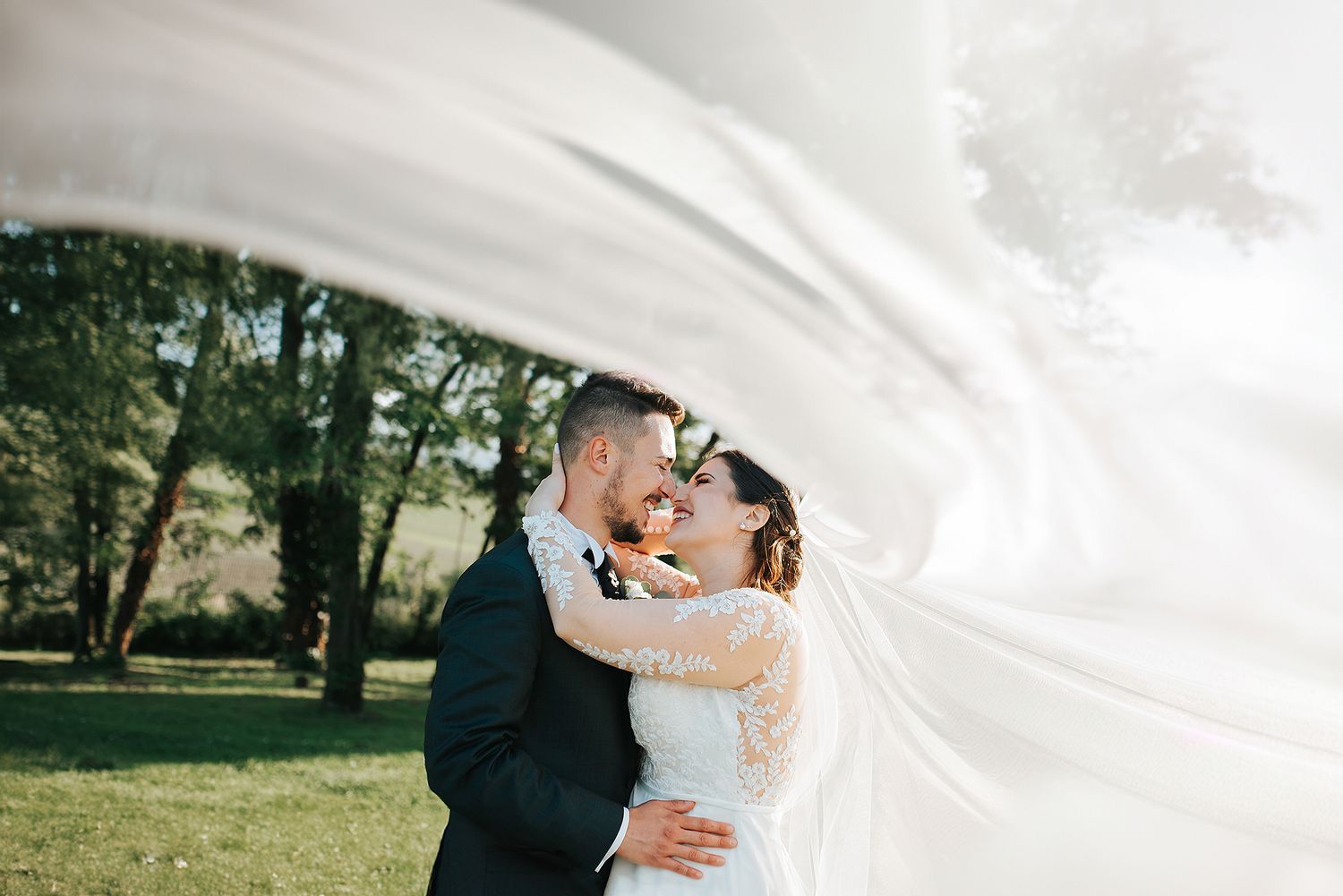 Brides veil billows dramatically as couple shares a kiss in a sunlit garden.