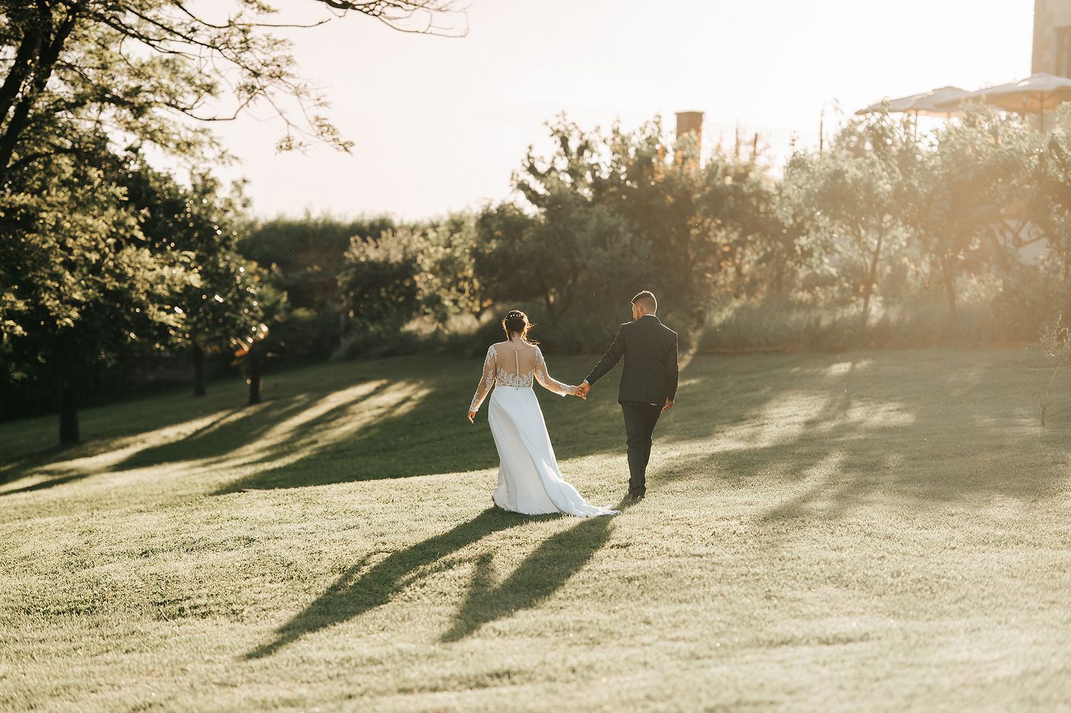 Bride and groom walking hand in hand across lush green lawn at golden hour sunset wedding.