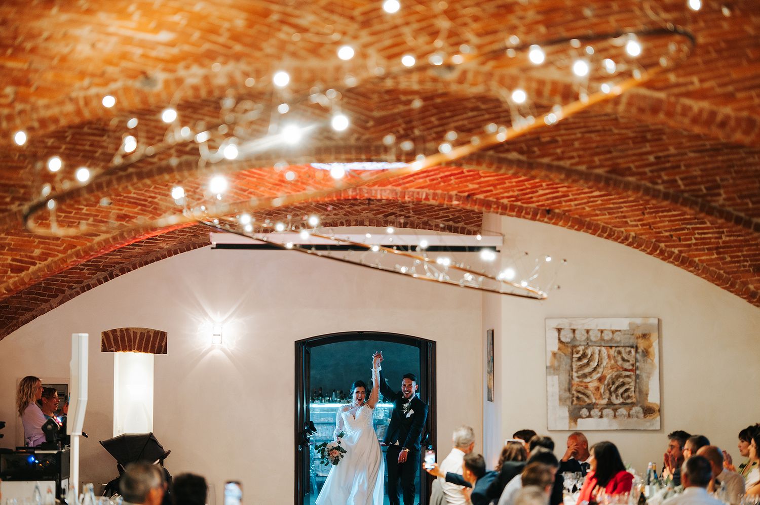 Bride and groom entering elegant rustic reception hall with brick arched ceiling and string lights.