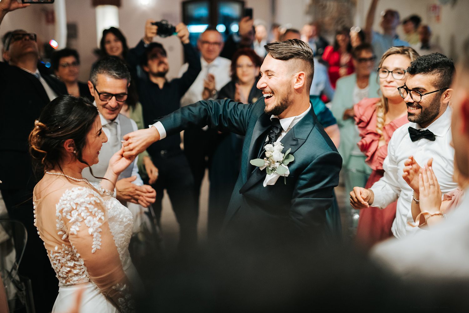 Joyful groom dancing with bride surrounded by cheering guests at lively wedding reception.