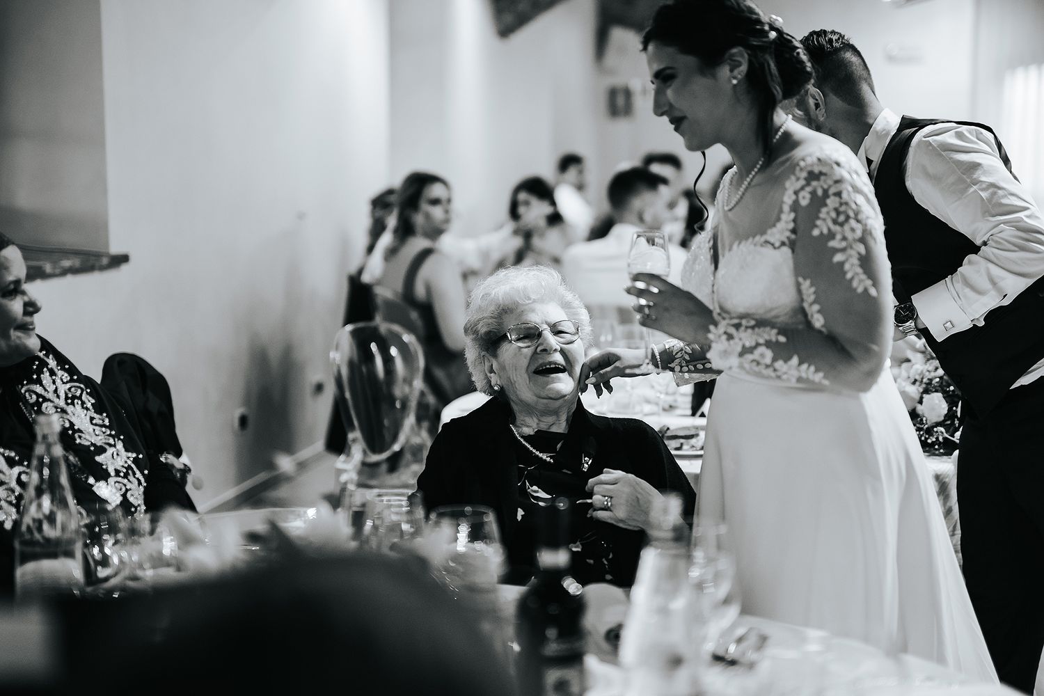 Bride in lace gown sharing warm moment with elderly guest at wedding reception table.
