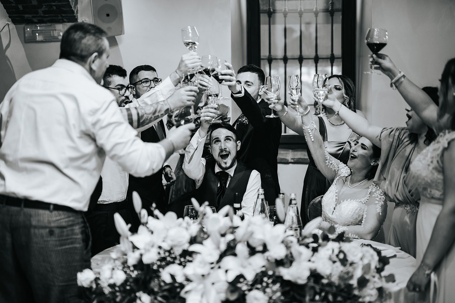 Wedding party raising glasses in joyful toast surrounded by floral centerpieces at reception.