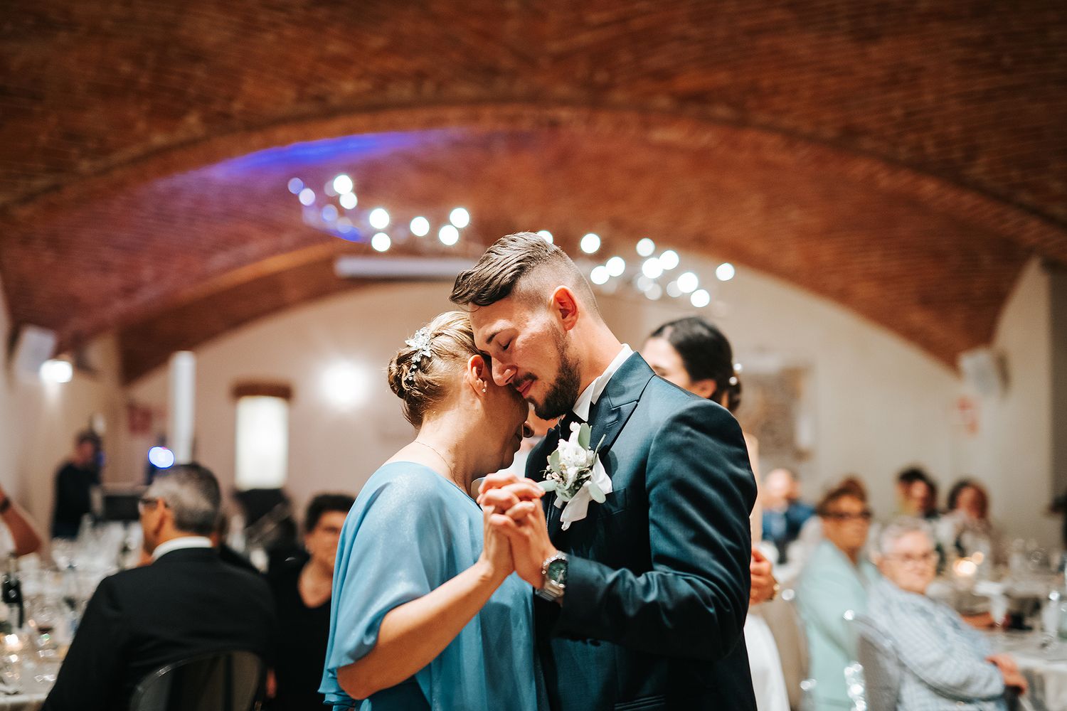 Couple shares tender first dance in rustic brick-vaulted wedding reception hall with string lights.