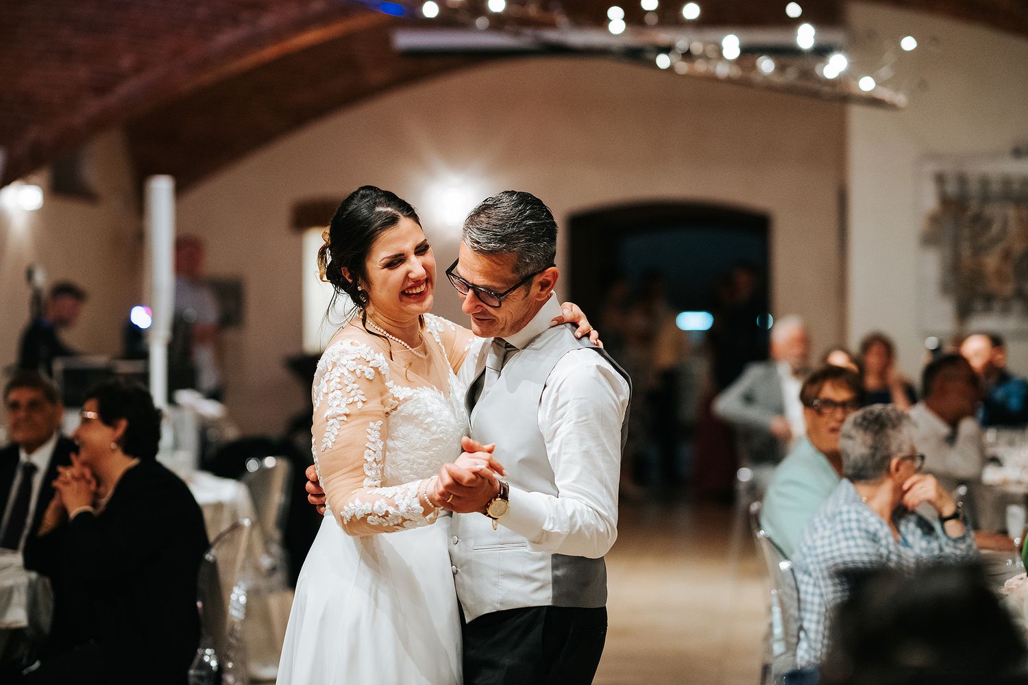 Bride in lace gown and groom dance joyfully at wedding reception in elegant vaulted ballroom.