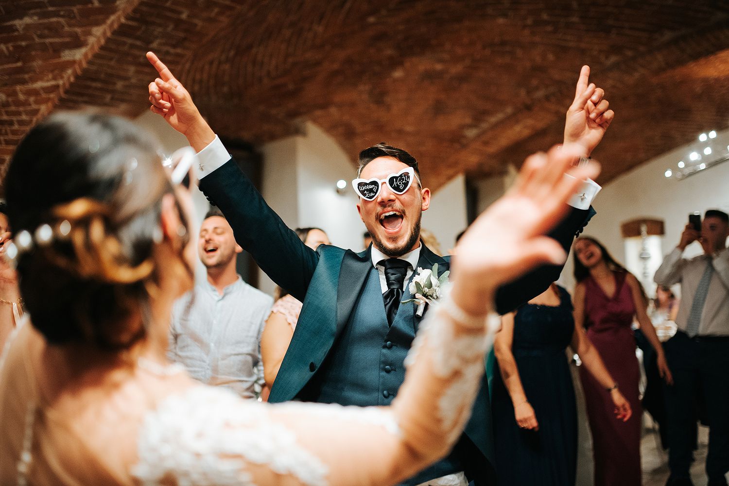 Groom in blue suit wearing heart sunglasses dancing joyfully at wedding reception party.