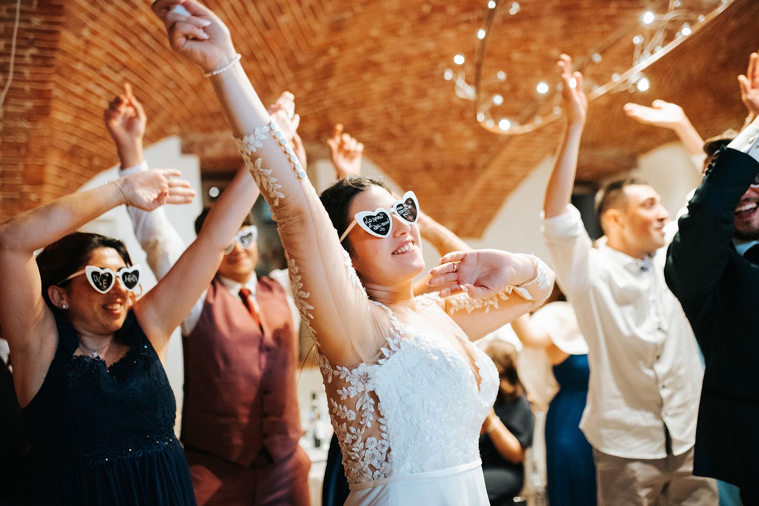 Bride and guests dancing joyfully wearing heart-shaped sunglasses at lively wedding reception.