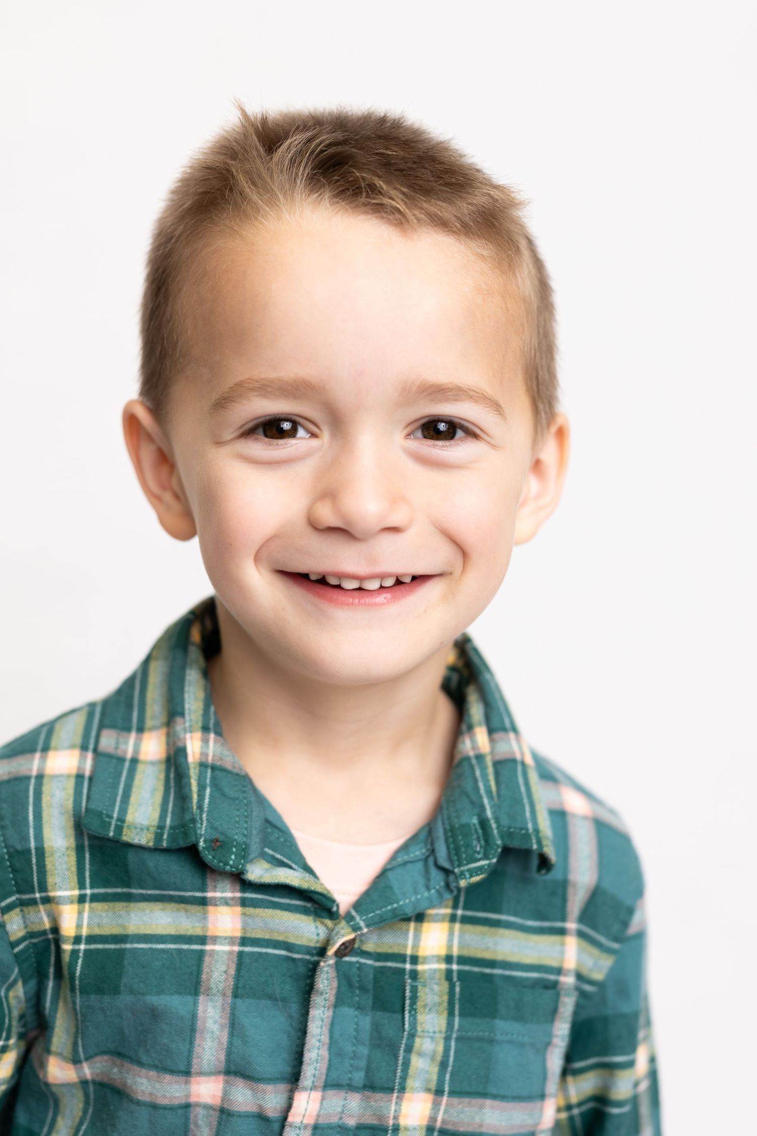 Young child in green plaid shirt showing big cheerful smile against white background.