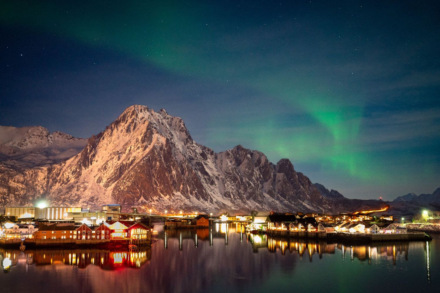 Northern lights illuminate a mountainous harbor town with glowing reflections on calm waters.