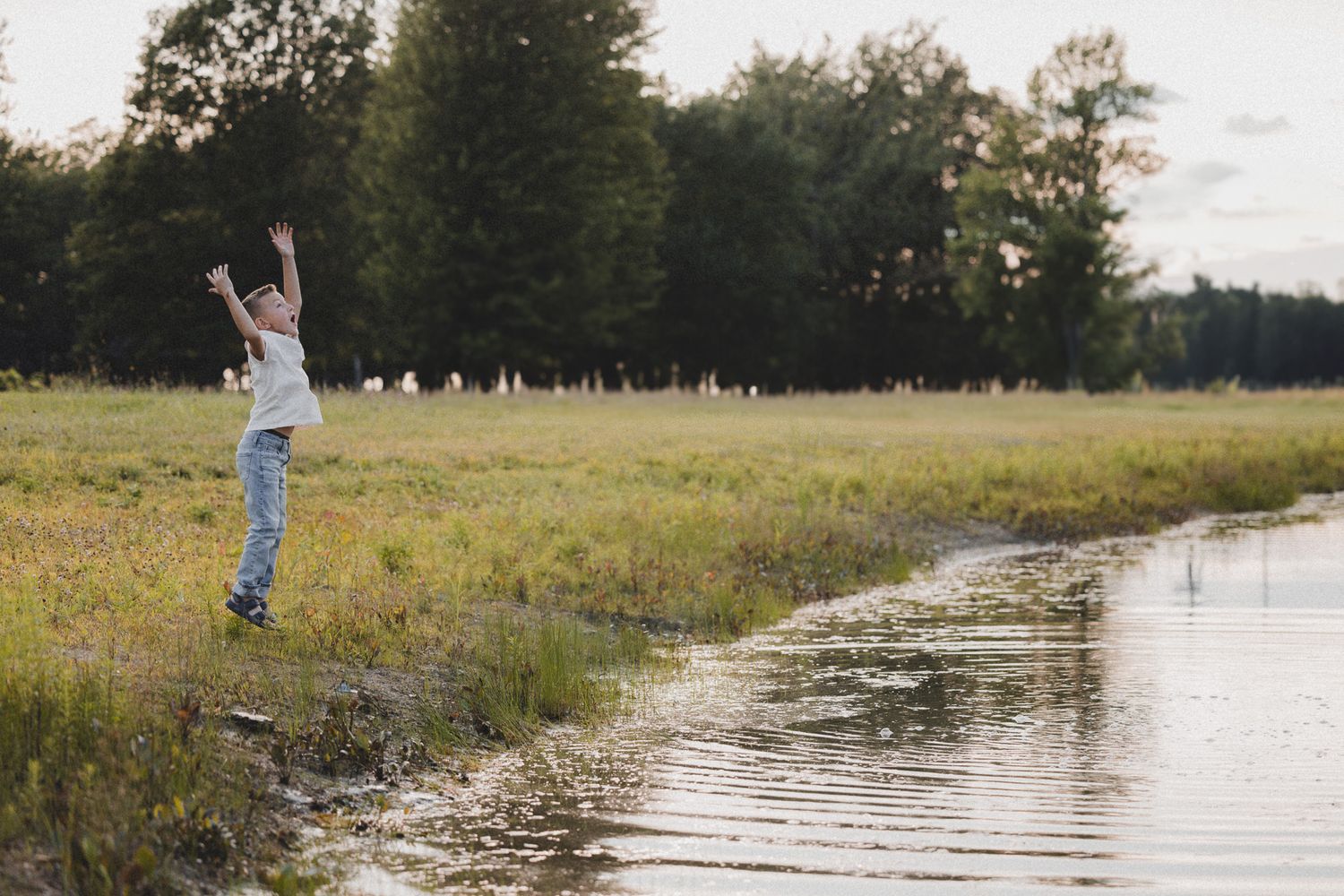 Person in jeans celebrates with arms raised beside a serene pond at golden hour.