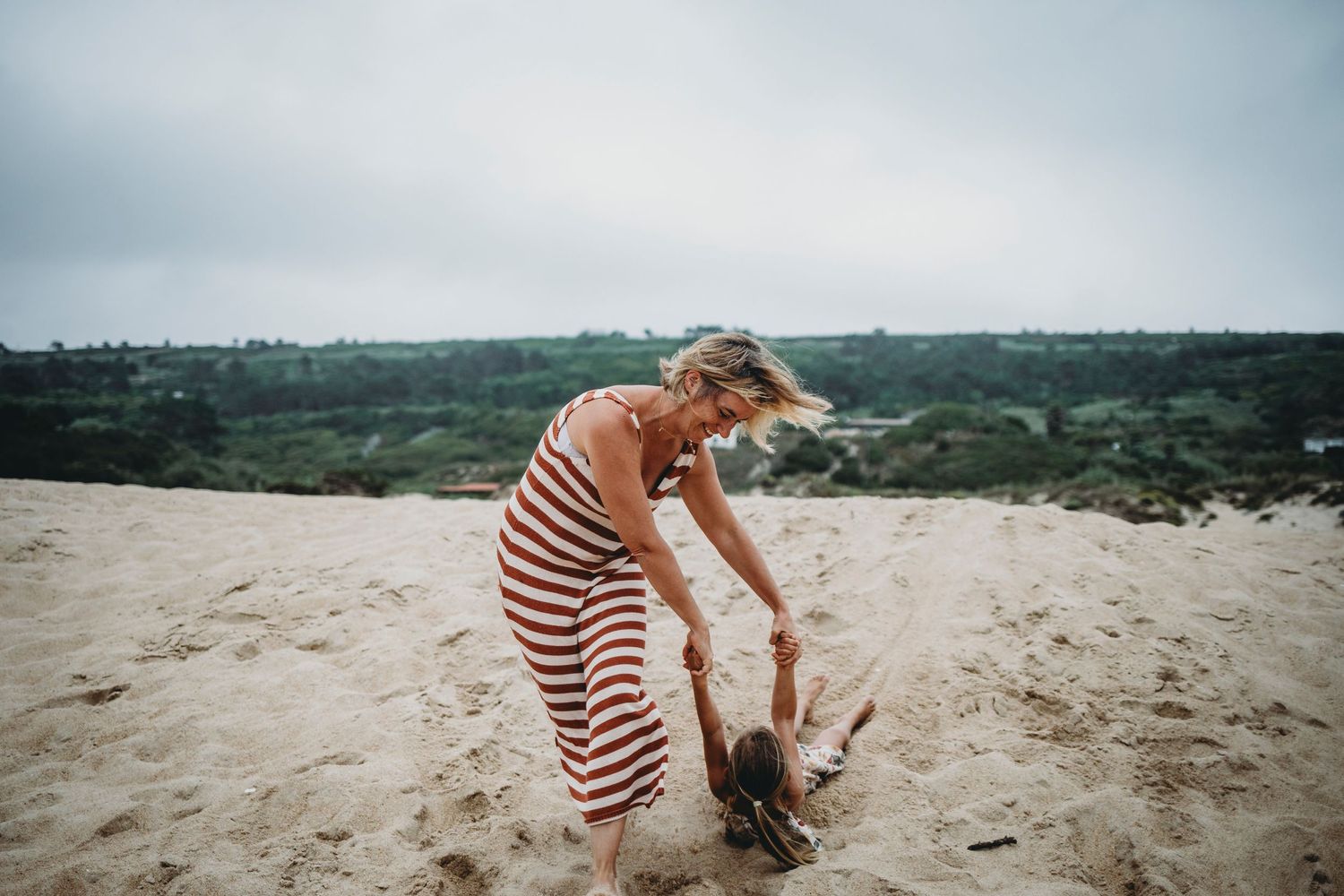 foto colorida na praia do Salgado, mãe brinca de arrastar a filha pela areia durante uma sessão fotográfica