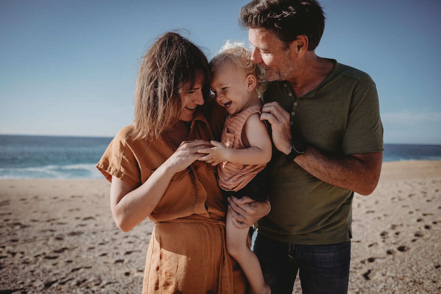 foto colorida pai, mãe e filho no colo com  na praia do Salgado durante sessão fotográfica de família