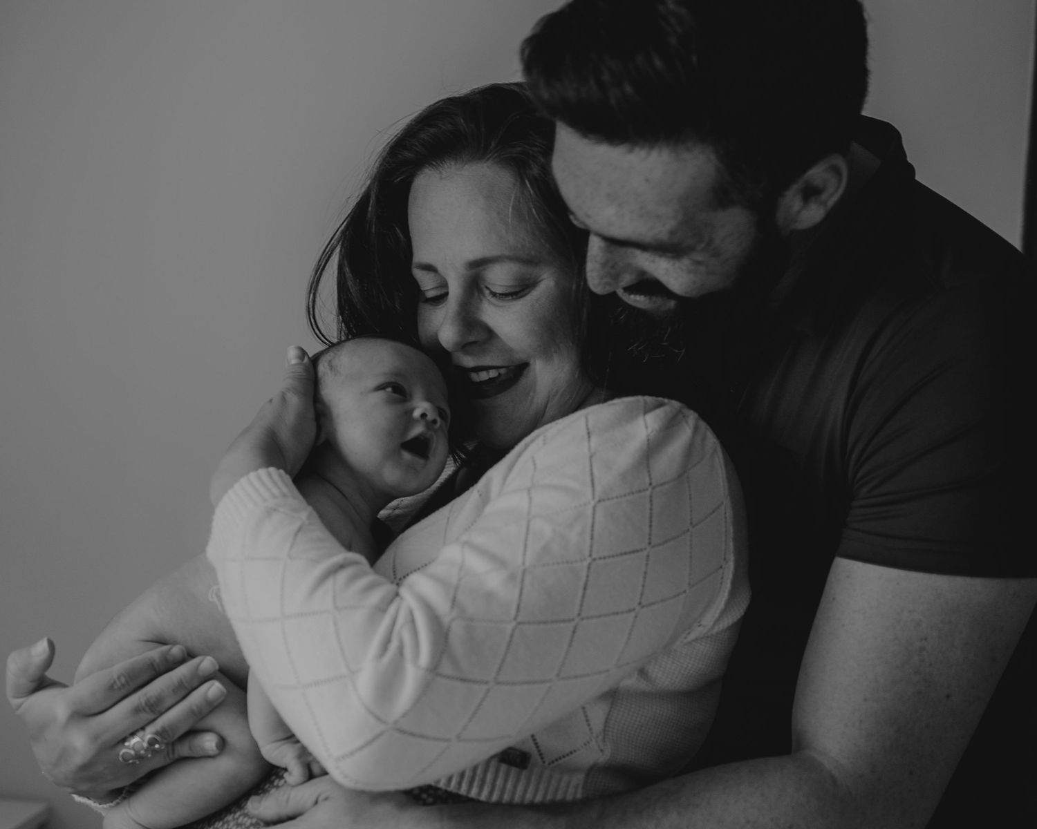 fotografia de família em preto e branco , o pai abraça a mãe e a mãe segura o bebé no colo sorrindo em fotografia família Caldas Da Rainha