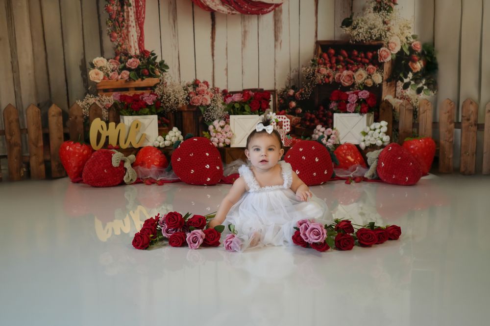 Baby in white dress sits on floor surrounded by red roses and Christmas presents against wooden fence backdrop.