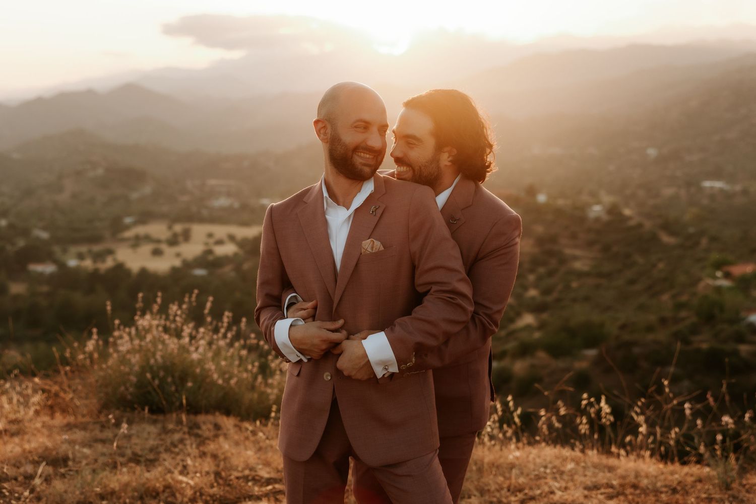 Two people embrace in rust-colored suits during golden hour on a mountain overlook.