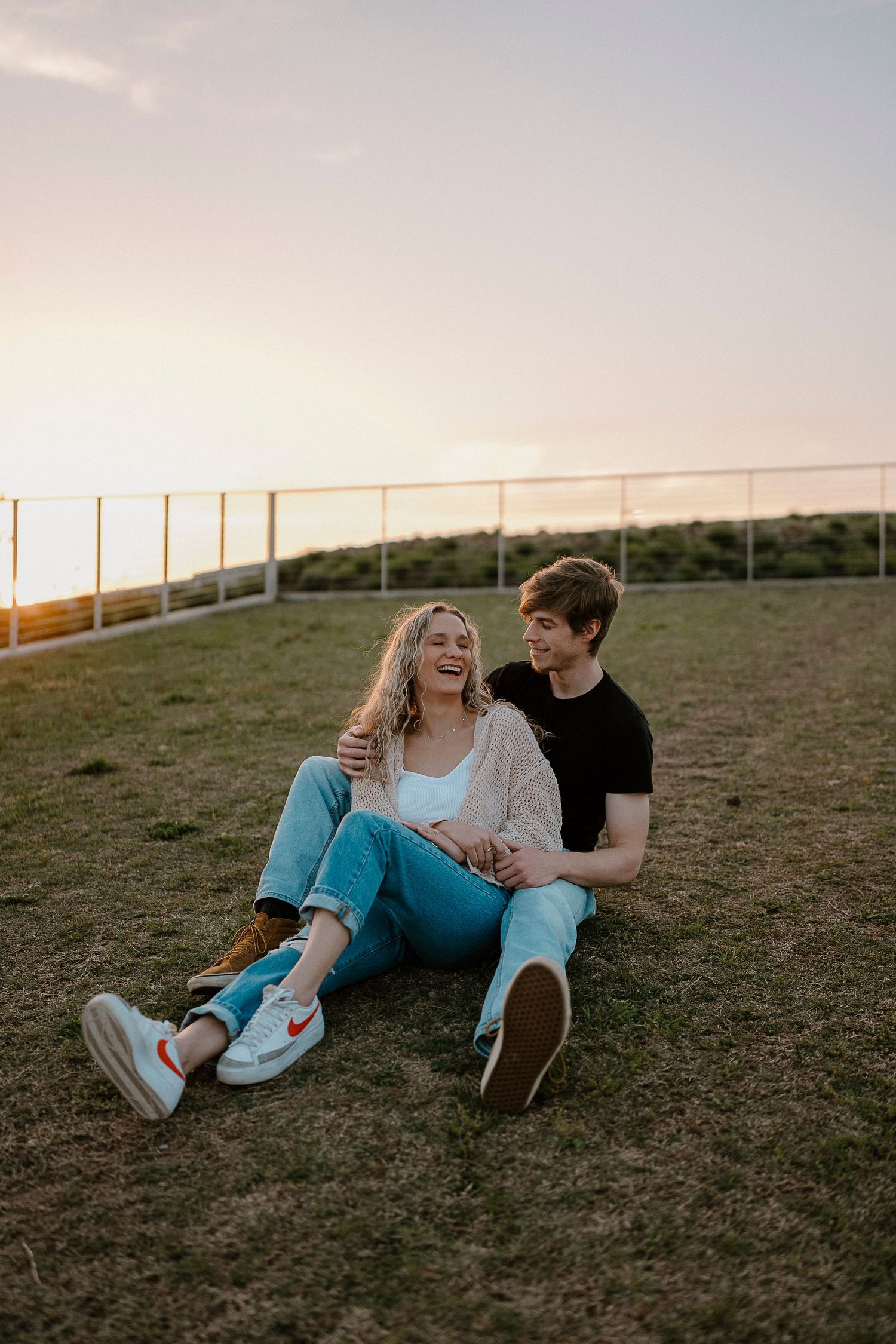 Two people share a laugh while sitting on grass during sunset.