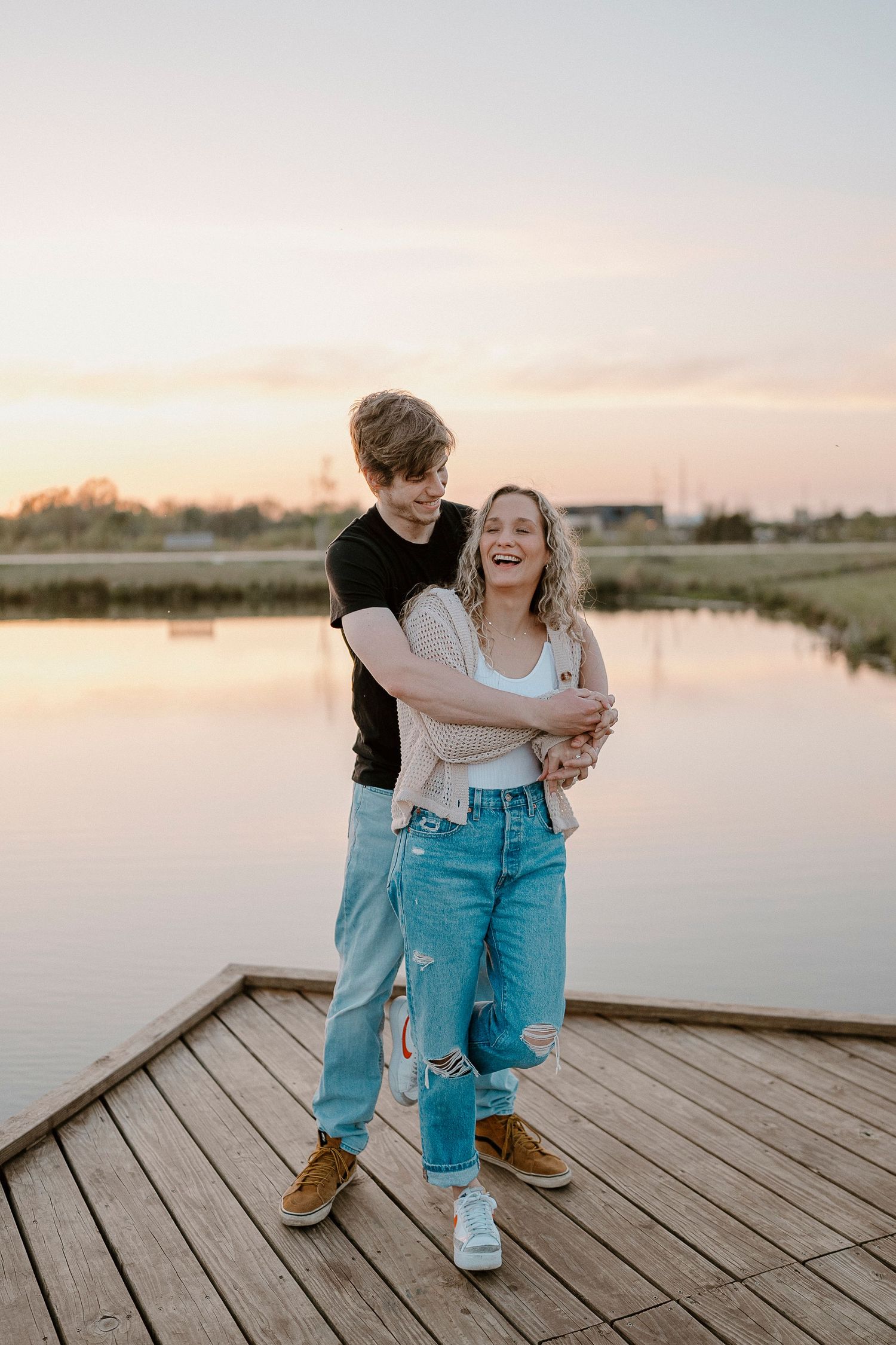 Two people in matching blue jeans share intimate moment on lakeside dock at dusk.