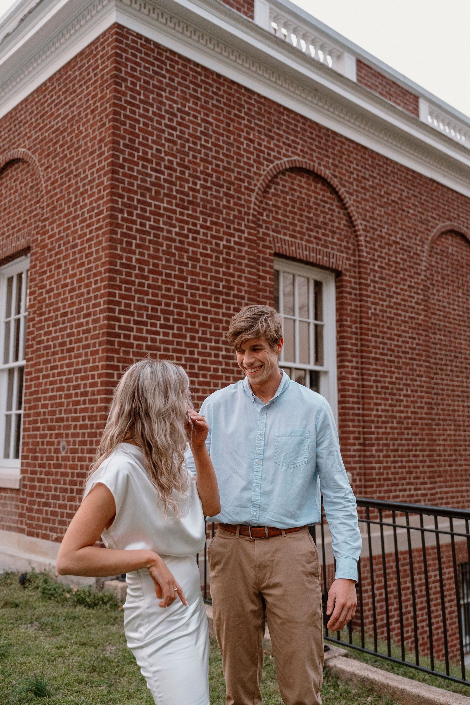 Couple shares playful moment outside traditional red brick building.