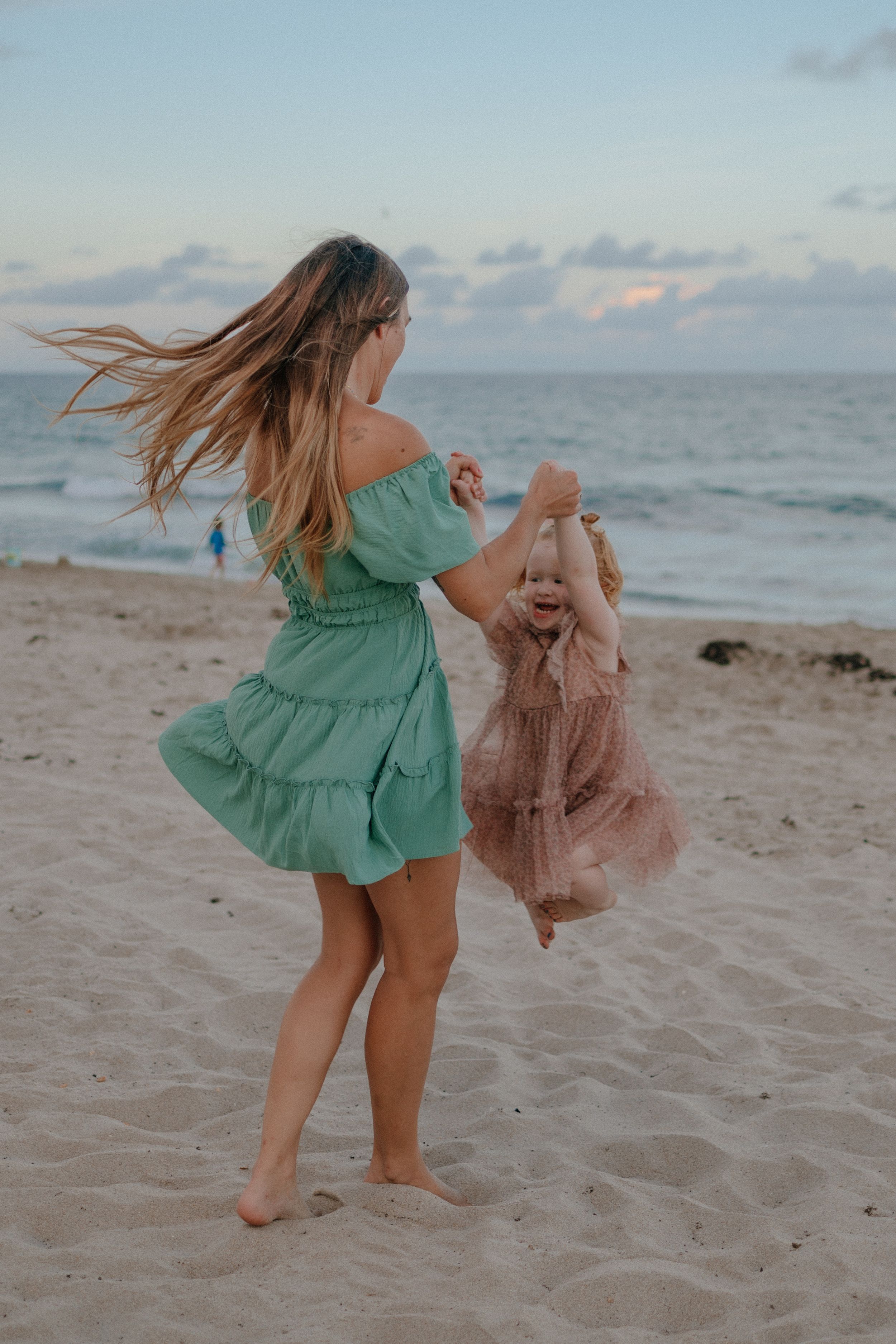 Woman in green dress twirling with small child in pink dress on sandy beach at sunset.