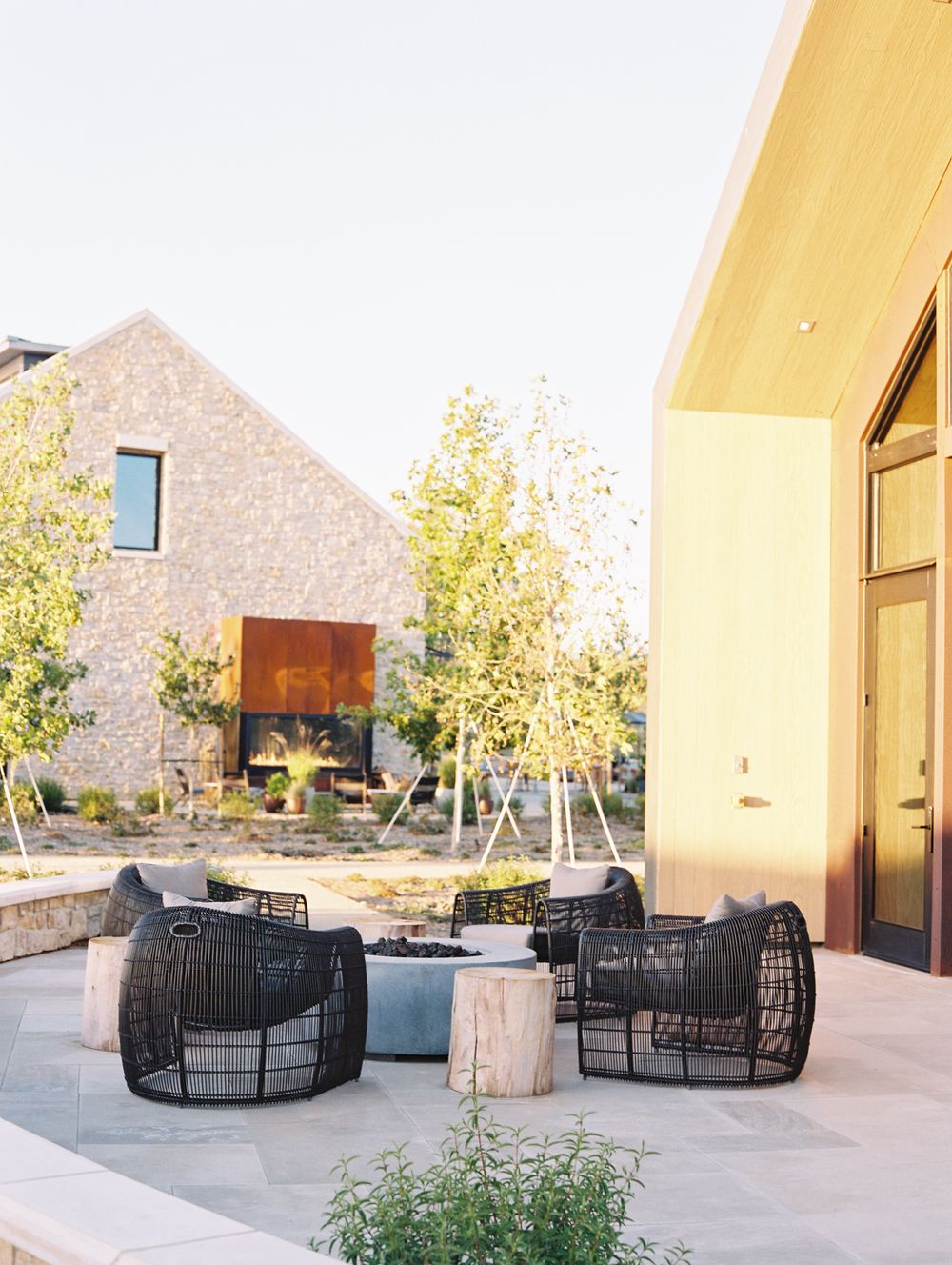 Modern winery patio with black woven chairs, fire pit, and stone building surrounded by young trees.