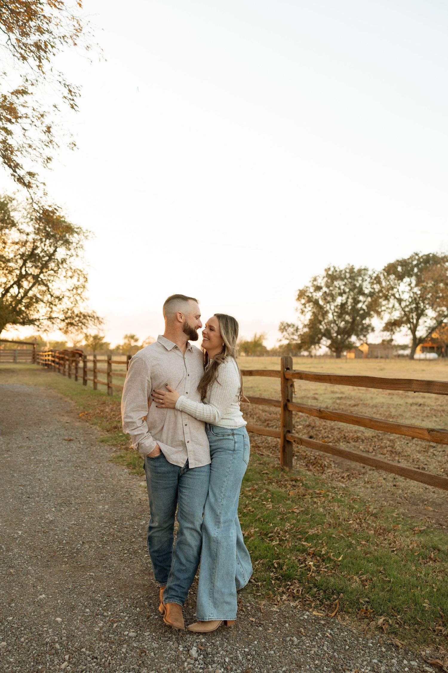 A couple embraces on a dirt path at sunset next to a wooden fence in a rustic countryside setting.