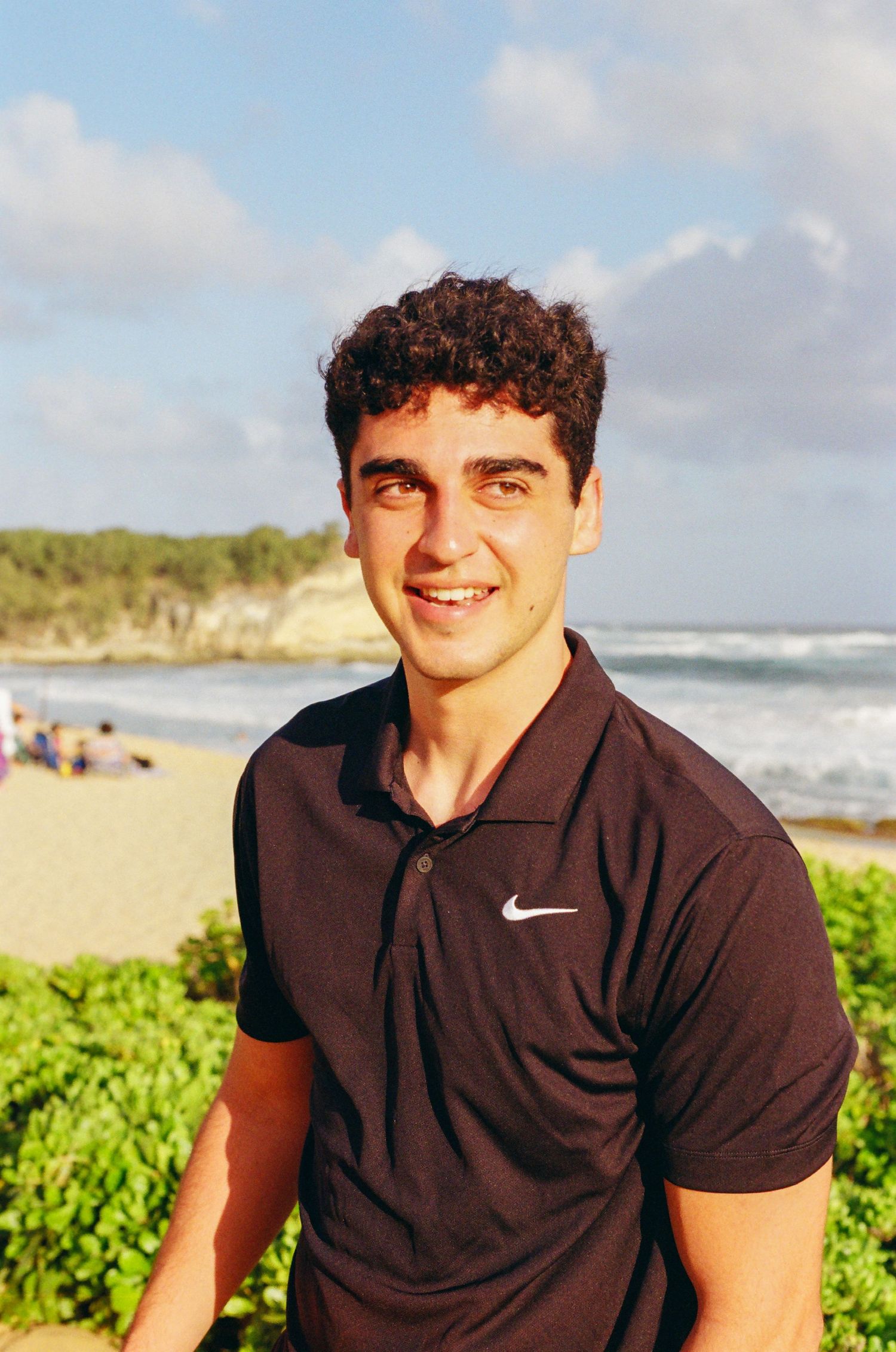 Young man in a black Nike polo shirt smiling at the beach with ocean waves and greenery in the background.