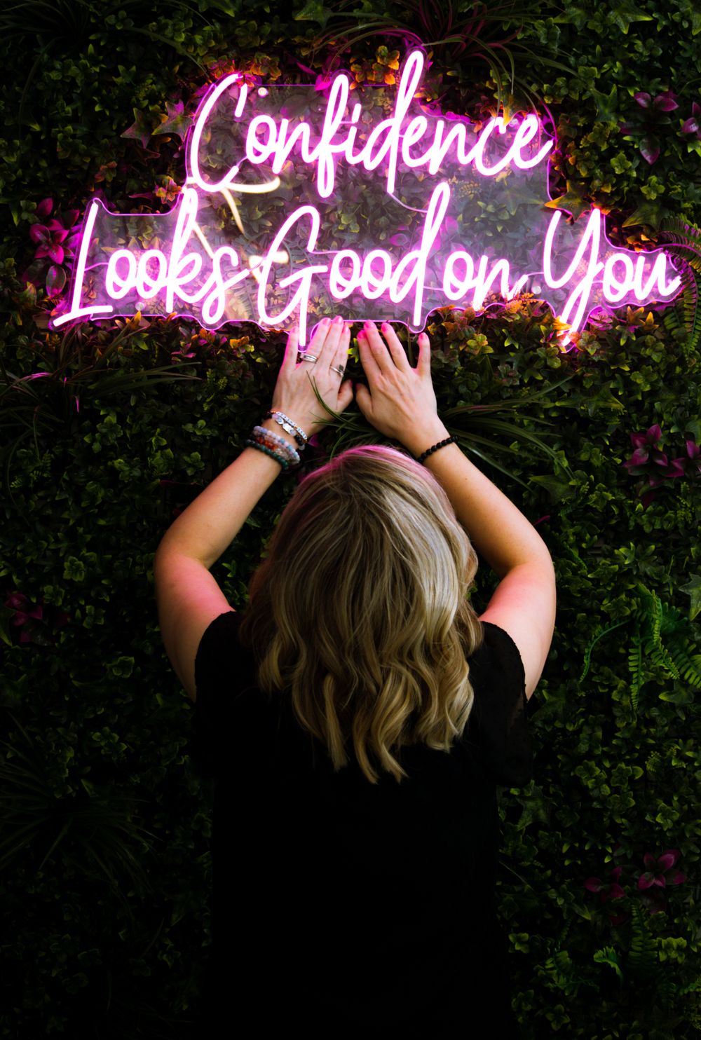 Pink neon sign reading 'Confidence Looks Good on You' glows against foliage.