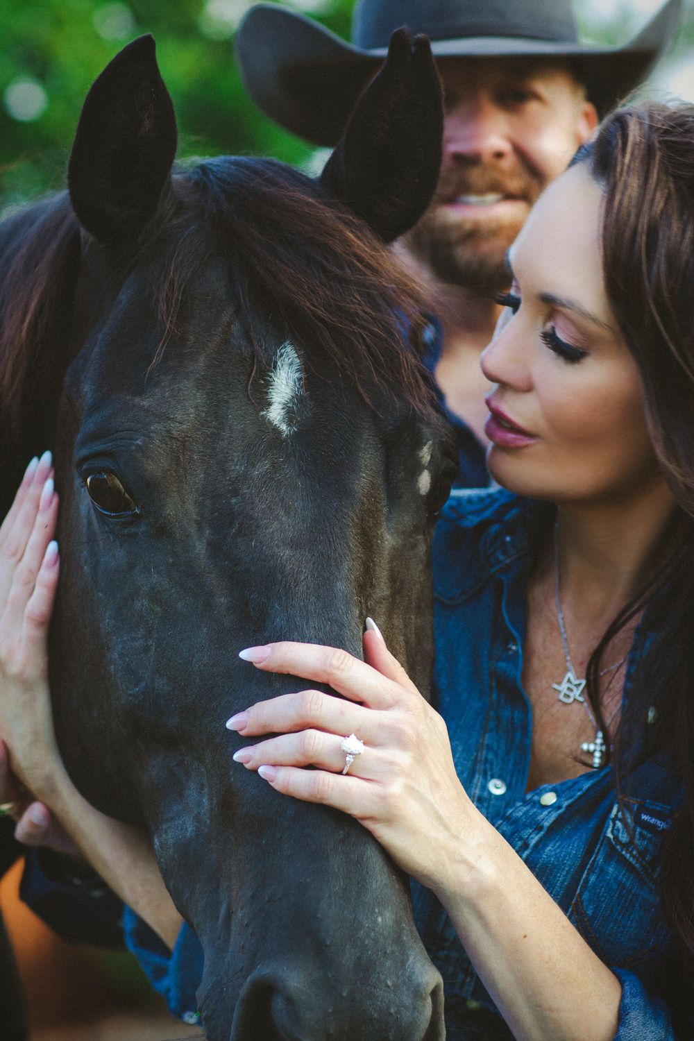 A black horse being gently petted while wearing a denim jacket and cowboy hat in a romantic country setting.