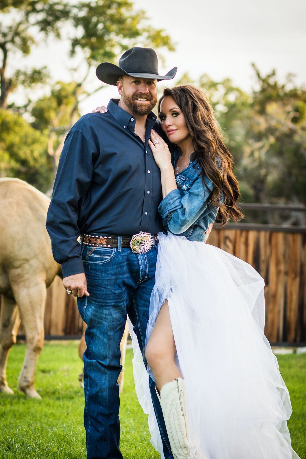 Country western couple in denim and formal wear pose in front of fence and horse.