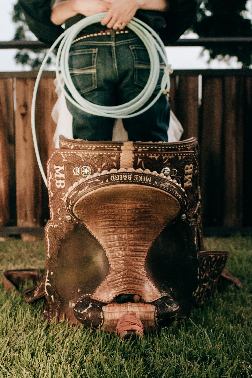 A vintage leather saddle and lasso rope displayed on grass against a wooden fence background.