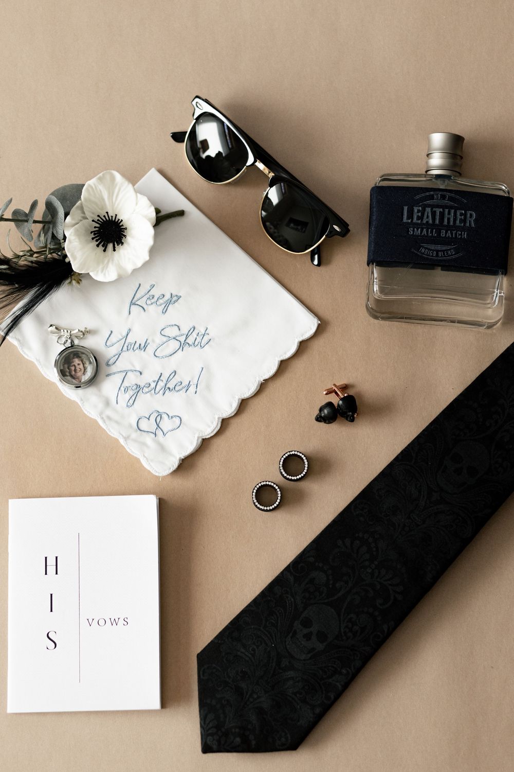 Flat lay of groom accessories including black tie, cologne, rings and handkerchief on beige background.