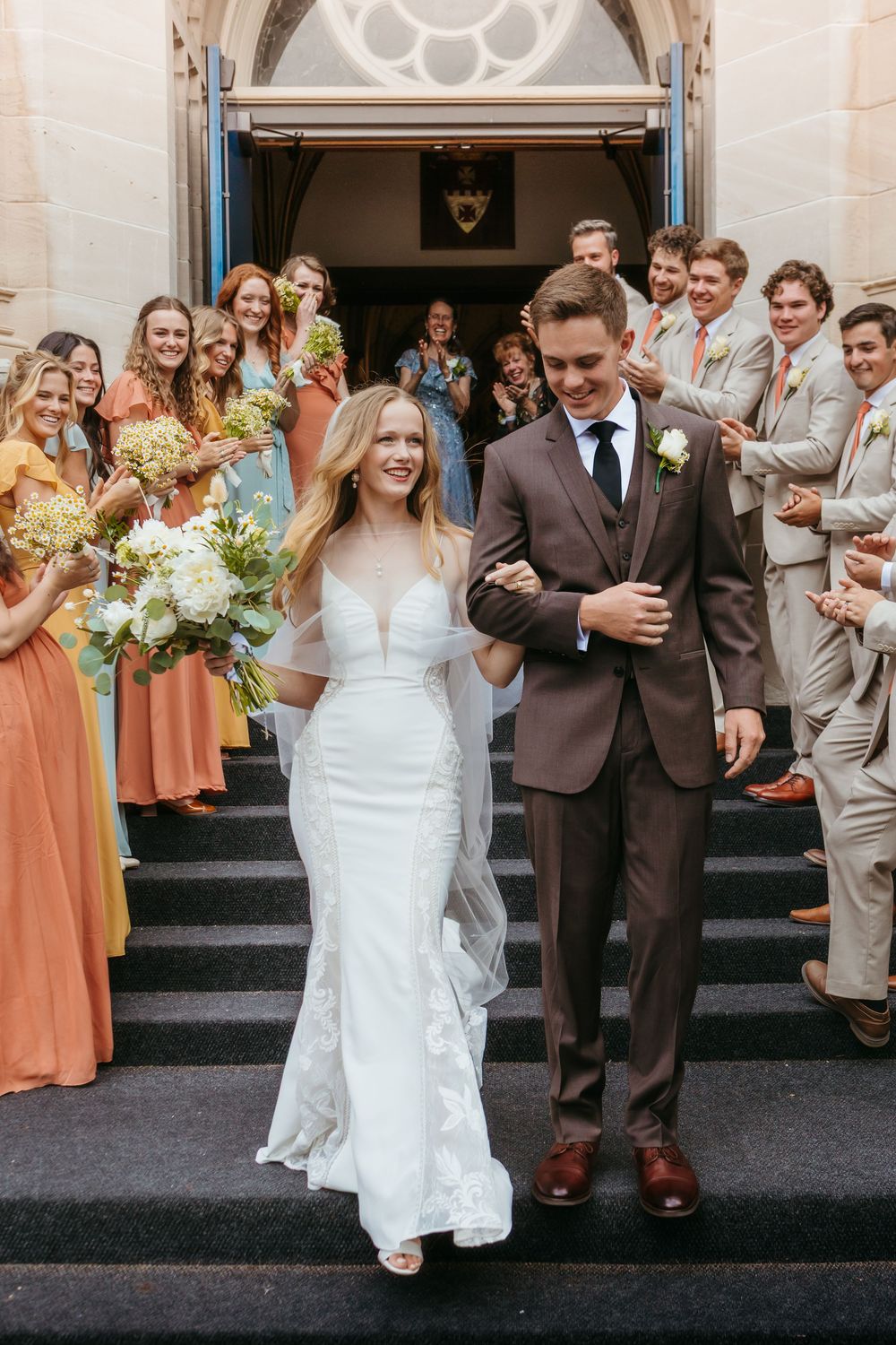 Wedding party celebration on church steps with bridesmaids in peach dresses and groomsmen in brown suits.