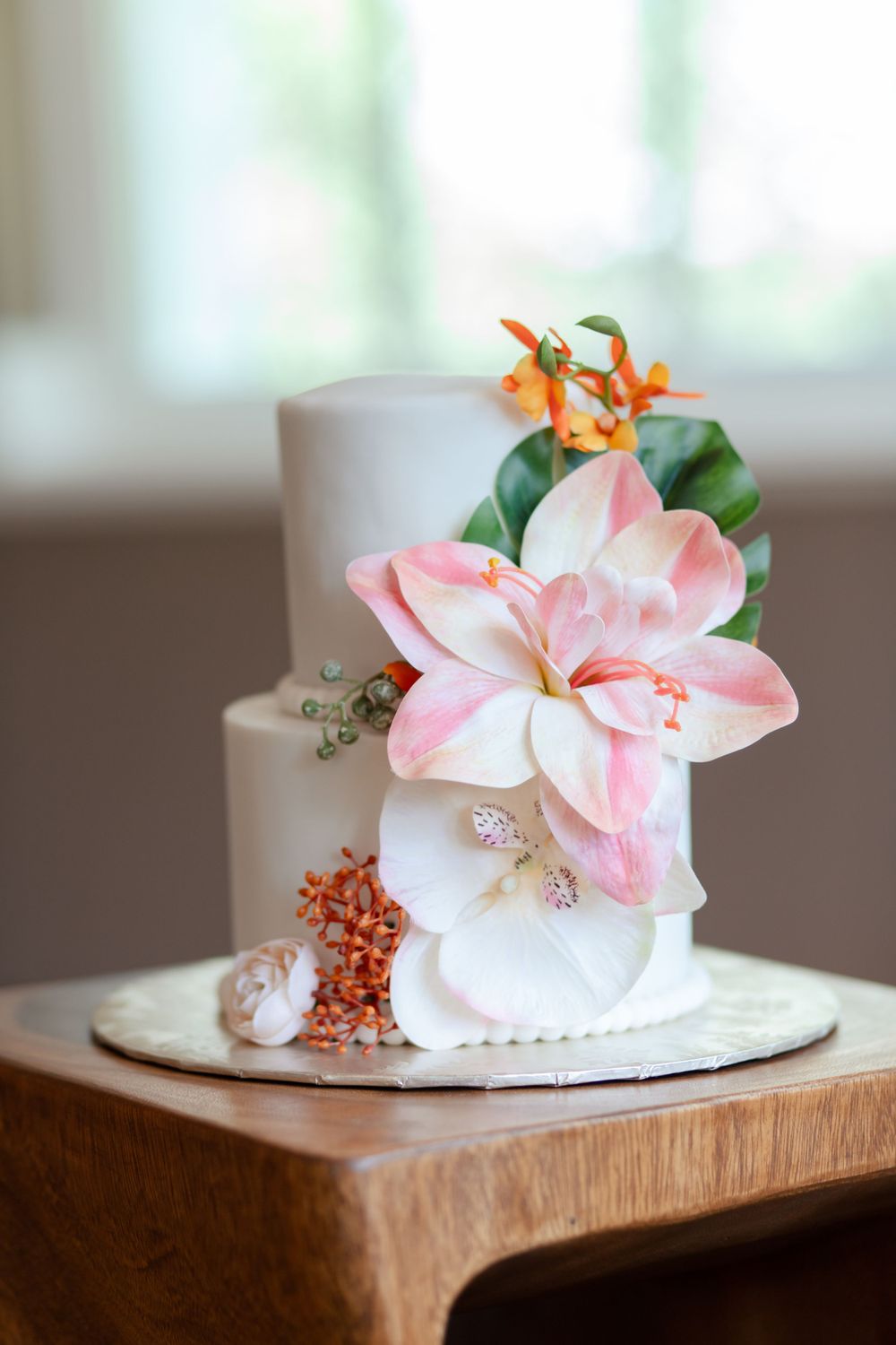A white two-tier wedding cake decorated with pink lilies and orange tropical flowers.
